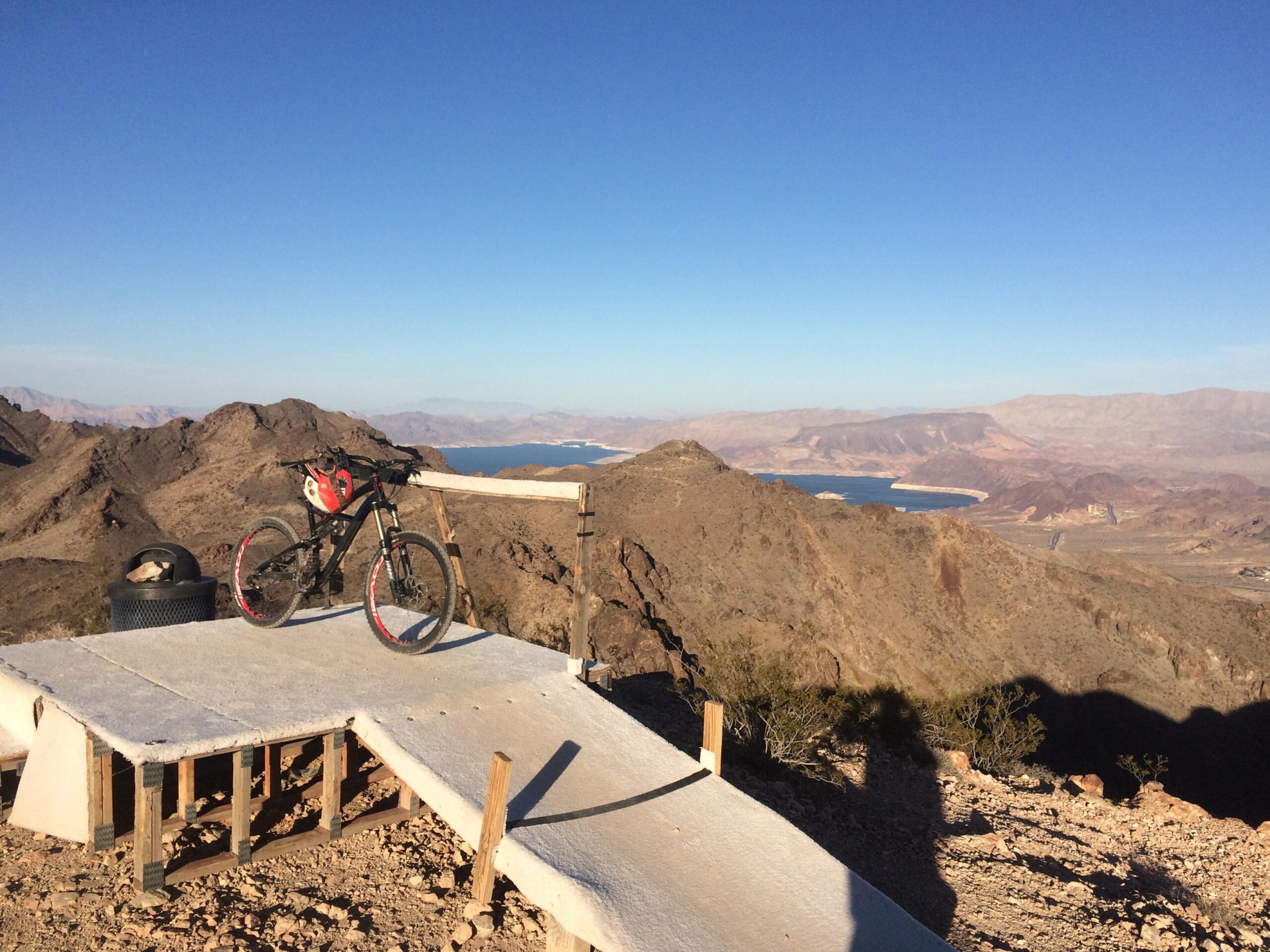 A mountain bike resting on a small wooden platform overlooking a scenic desert landscape. In the background, a blue lake is visible, surrounded by rocky mountains under a clear blue sky. A trash can sits on the platform, adding to the outdoor setting. Bootleg Canyon mountain bike trail.