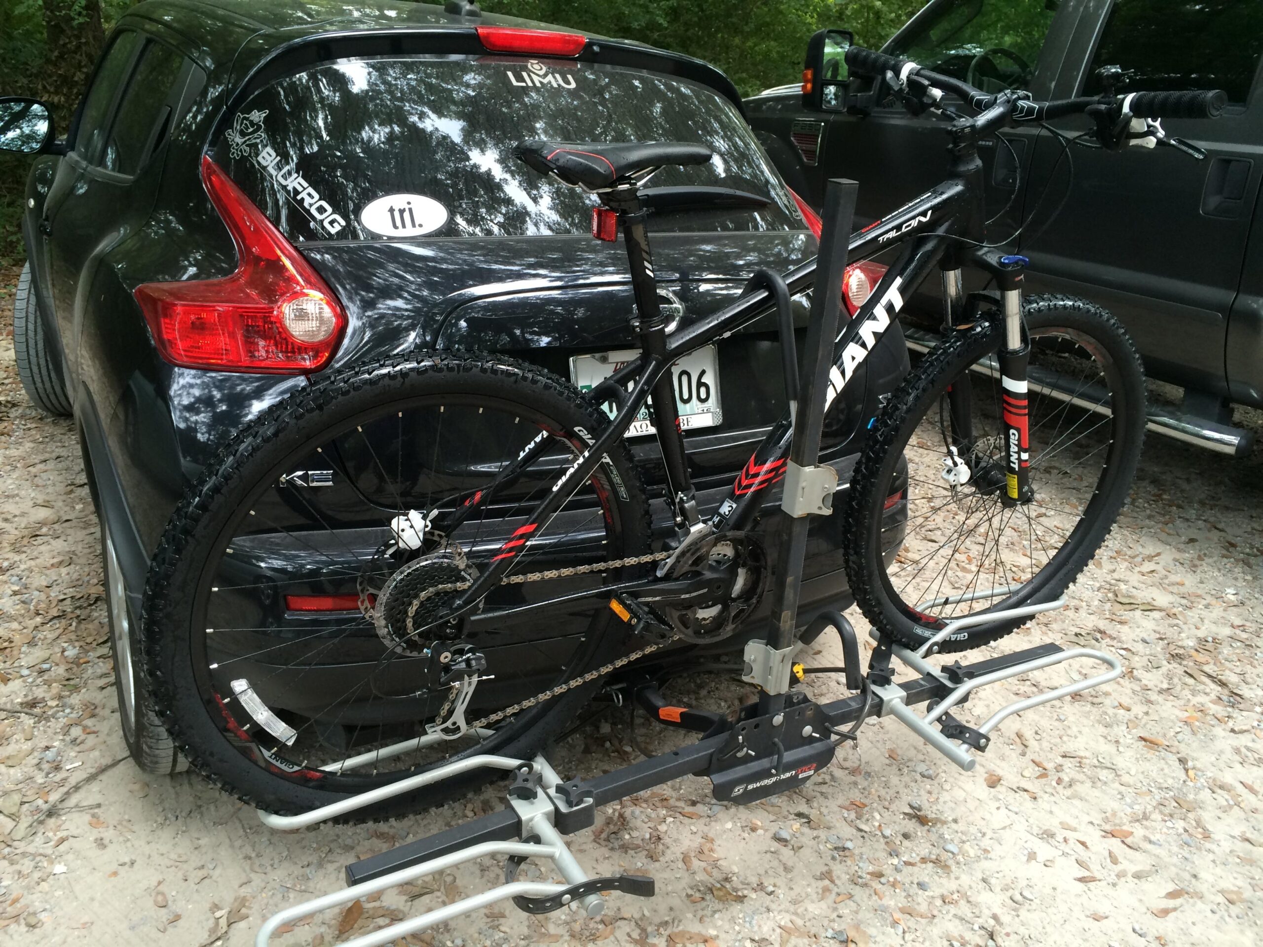 Giant Talon 27.5: A black car parked on a gravel surface with a mountain bike mounted on a hitch rack at the back. The bike features a dark frame and thick tires, and nearby stickers are visible on the car's rear window. Surrounding flora includes trees and foliage.