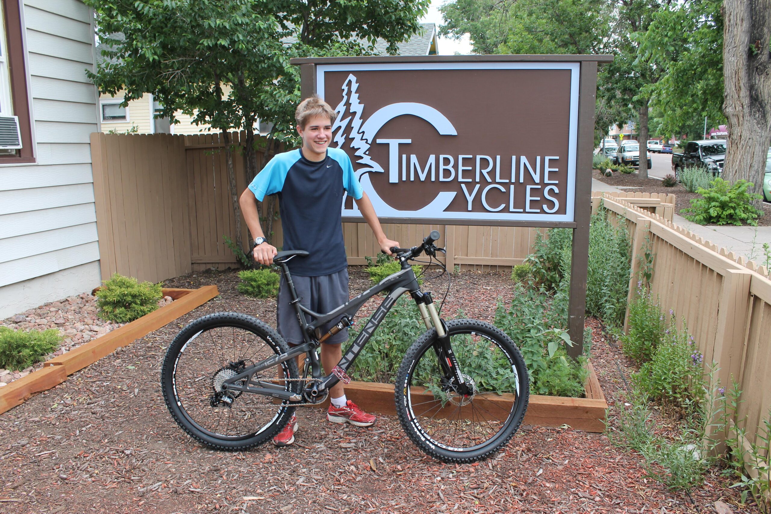 A young person standing beside a mountain bike in front of a sign for Timberline Cycles. The background features a landscaped area with small plants and a wooden fence. The individual is wearing a blue and black shirt, gray shorts, and red shoes, smiling at the camera.