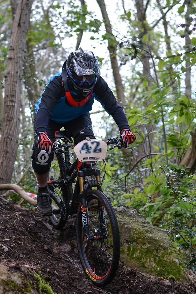 A mountain biker dressed in protective gear navigates a steep, wooded trail. The cyclist is wearing a full-face helmet, gloves, and a jersey with bright colors, focusing intently as they descend among trees and rocky terrain. The bike features racing number 42, emphasizing the competitive nature of the scene. St Brelade loop smash mountain bike trail.