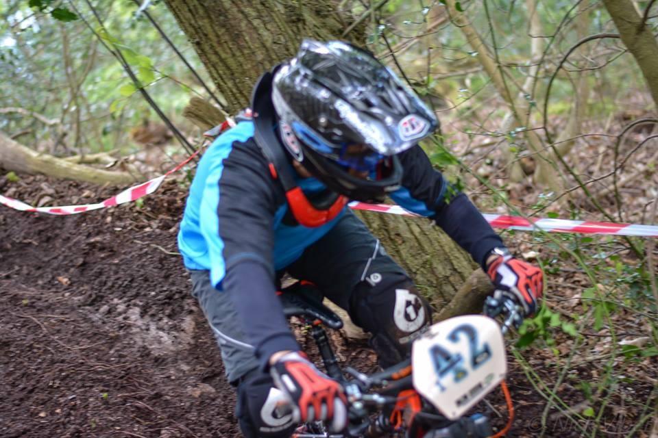 A mountain biker wearing a helmet and protective gear navigates a narrow, muddy trail in a wooded area, with red and white tape marking the course. The biker's focus is on the challenging terrain ahead. St Brelade loop smash mountain bike trail.