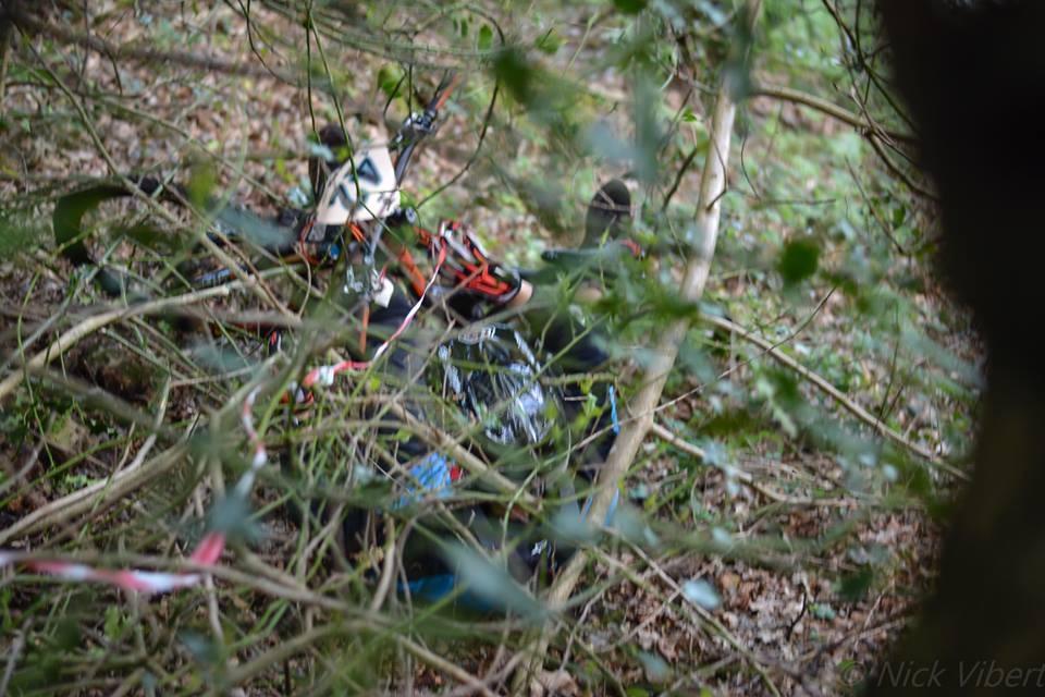 A partially obscured view of a person in a forested area, lying on the ground among underbrush and fallen branches. The individual appears to be wearing a black outfit with colorful accents. St Brelade loop smash mountain bike trail.
