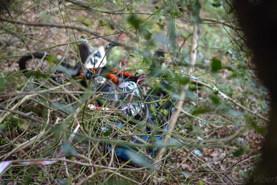 A partially obscured off-road motorcycle lies on the ground, surrounded by branches and foliage in a wooded area. St Brelade loop smash mountain bike trail.
