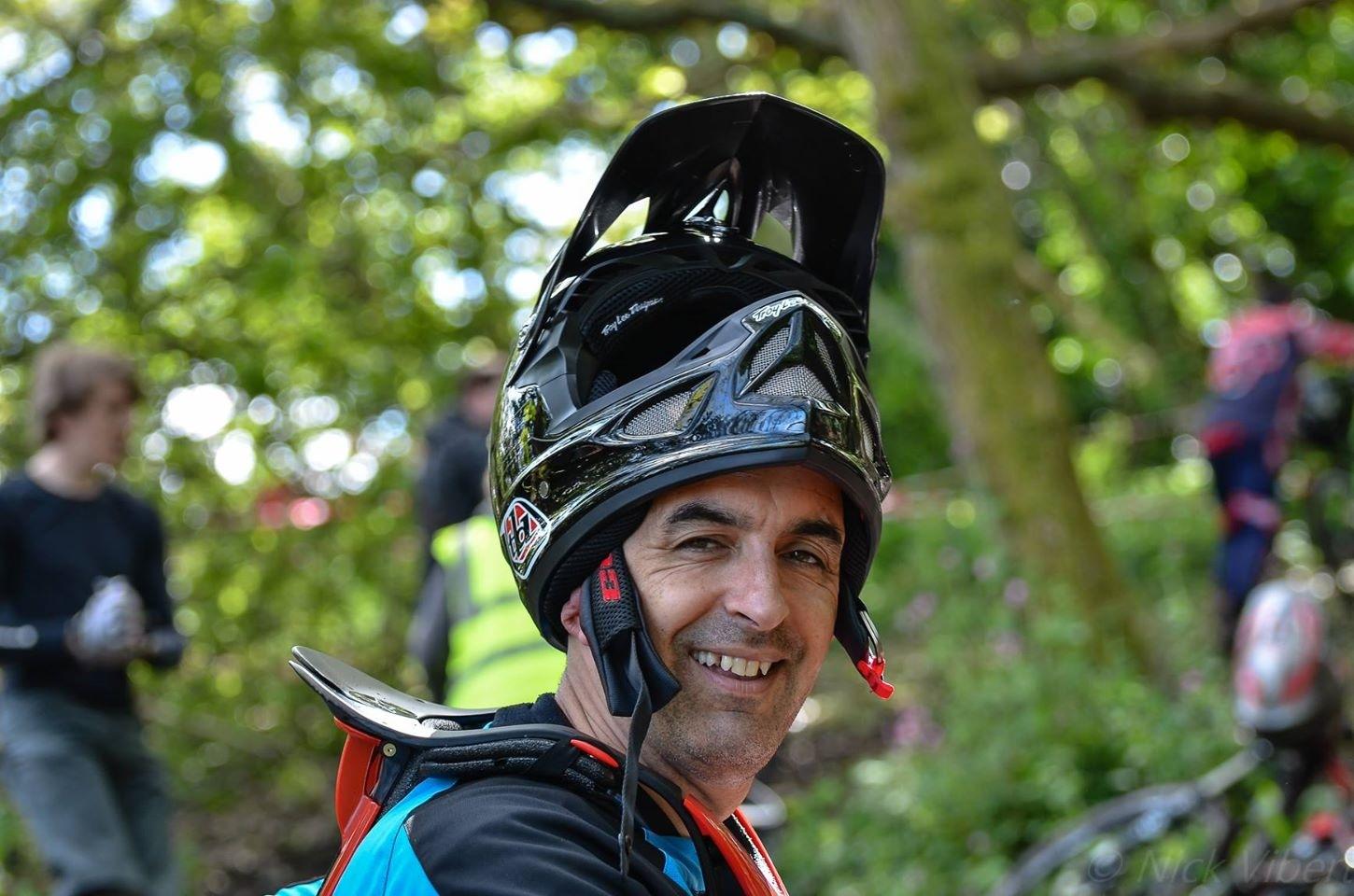 A smiling man wearing a black motocross helmet and protective gear, sitting in a green outdoor setting with blurred figures in the background, engaged in a motocross event. St Brelade loop smash mountain bike trail.