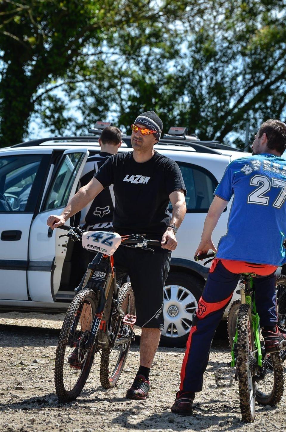 Two men in casual sports attire are chatting while standing next to their mountain bikes in a parking area. One man, wearing a black t-shirt with "LAZER" printed on it and sunglasses, is leaning on his bike. The other man, wearing a blue shirt with "SPEED" and the number 27, is also on a bike. A white car and a forested area can be seen in the background, under a clear blue sky. St Brelade loop smash mountain bike trail.