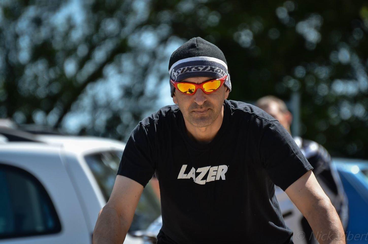 A man wearing sunglasses and a black beanie, with a t-shirt that reads "LAZER," is seen in an outdoor setting. He appears focused and determined, with blurred backgrounds of vehicles and trees. Sunlight illuminates the scene, highlighting the man's serious expression. St Brelade loop smash mountain bike trail.