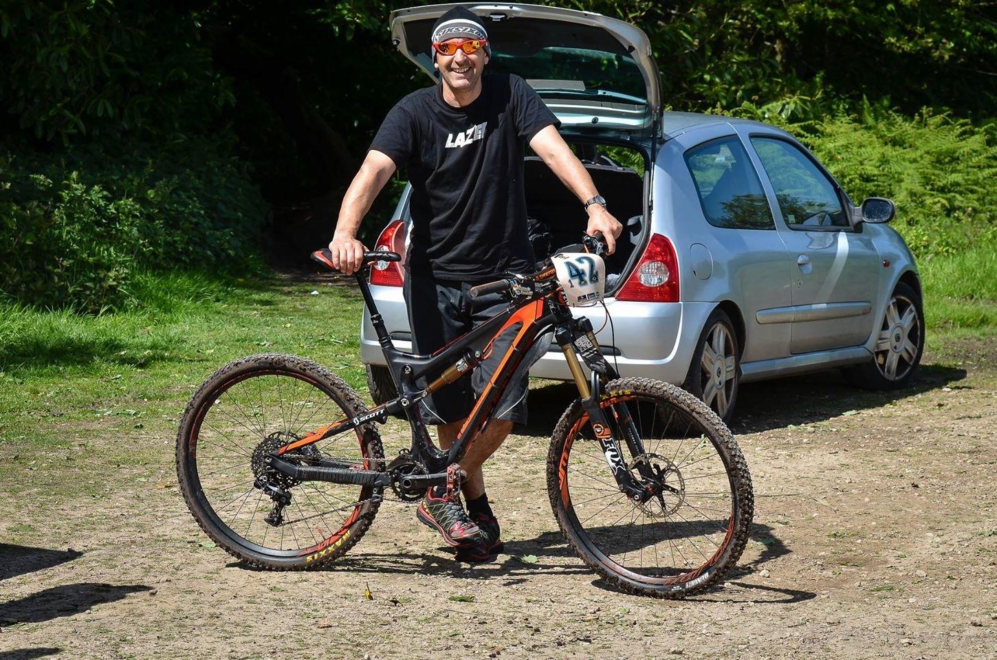 A smiling man wearing a black t-shirt and shorts stands next to an orange and black mountain bike. He holds the handlebars while resting one foot on the ground. The background features a silver car with its trunk open and green foliage. The scene is set on a sunny day in a grassy area. The bike has a race number displayed on it. St Brelade loop smash mountain bike trail.