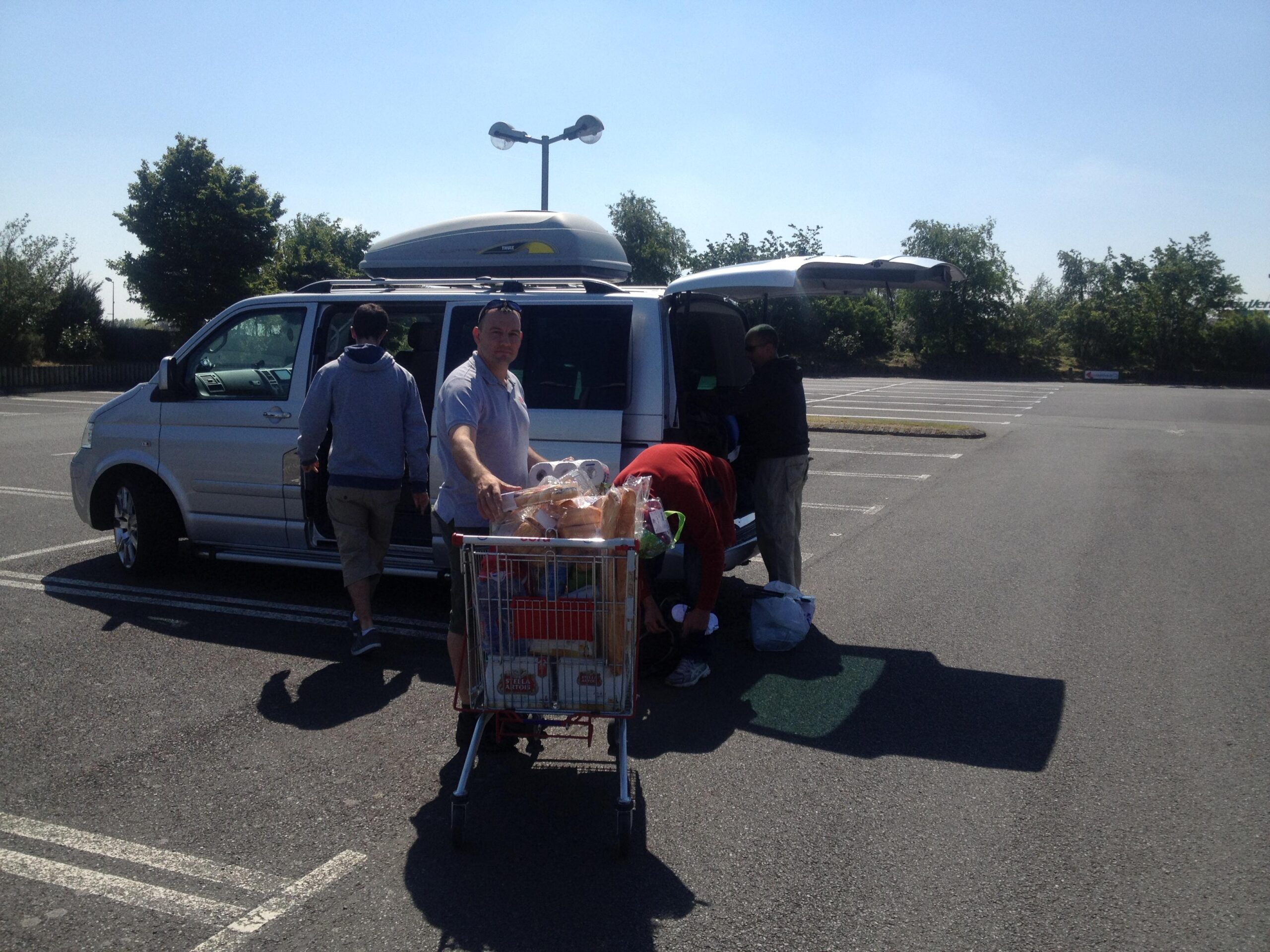 A silver van parked in an empty lot on a sunny day, with three people unloading groceries from a shopping cart. One person is pushing the cart while another is taking items from the back of the van. Trees and a clear blue sky are visible in the background. St Brelade loop smash mountain bike trail.