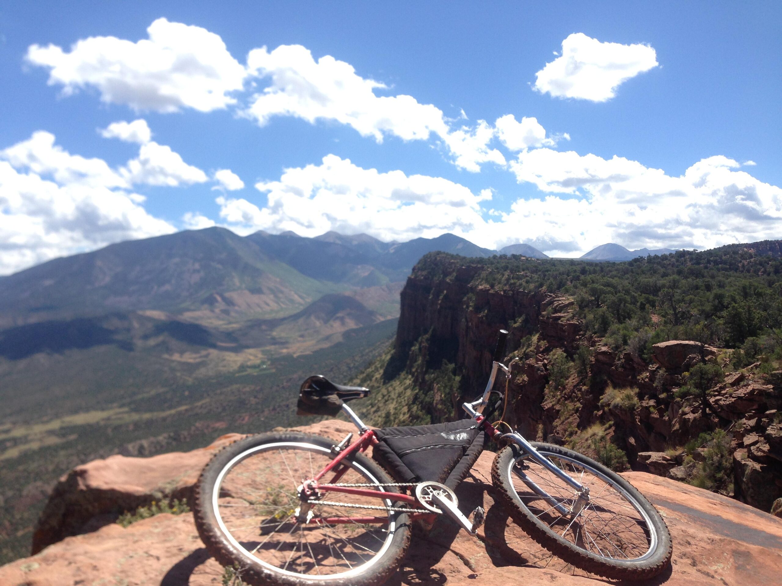 Trek 990: A mountain bike resting on a rocky ledge, overlooking a vast mountainous landscape with lush greenery and distant peaks under a blue sky adorned with fluffy white clouds.
