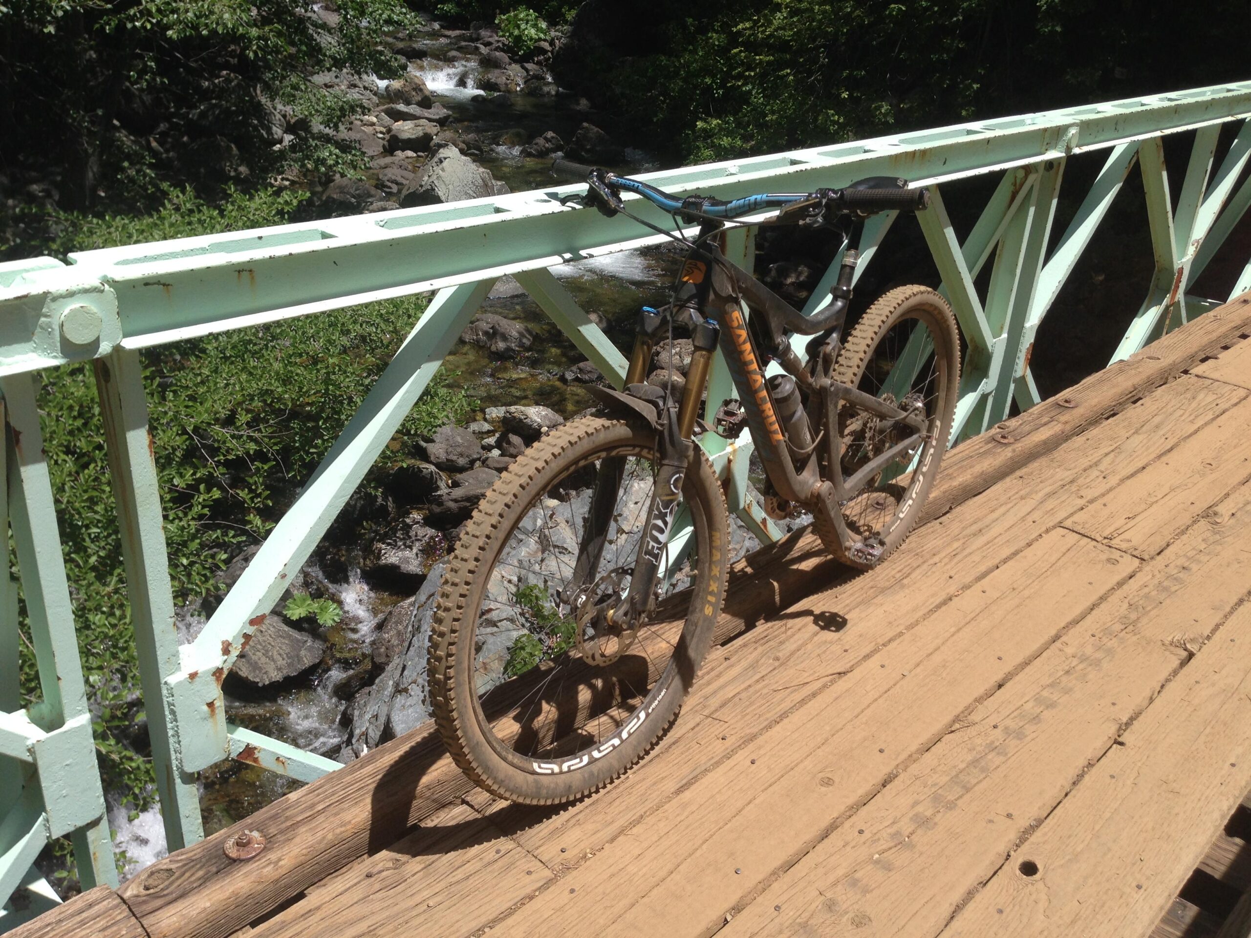 Santa Cruz Bronson: Mountain bike resting on a wooden bridge overlooking a rocky stream, surrounded by lush greenery.