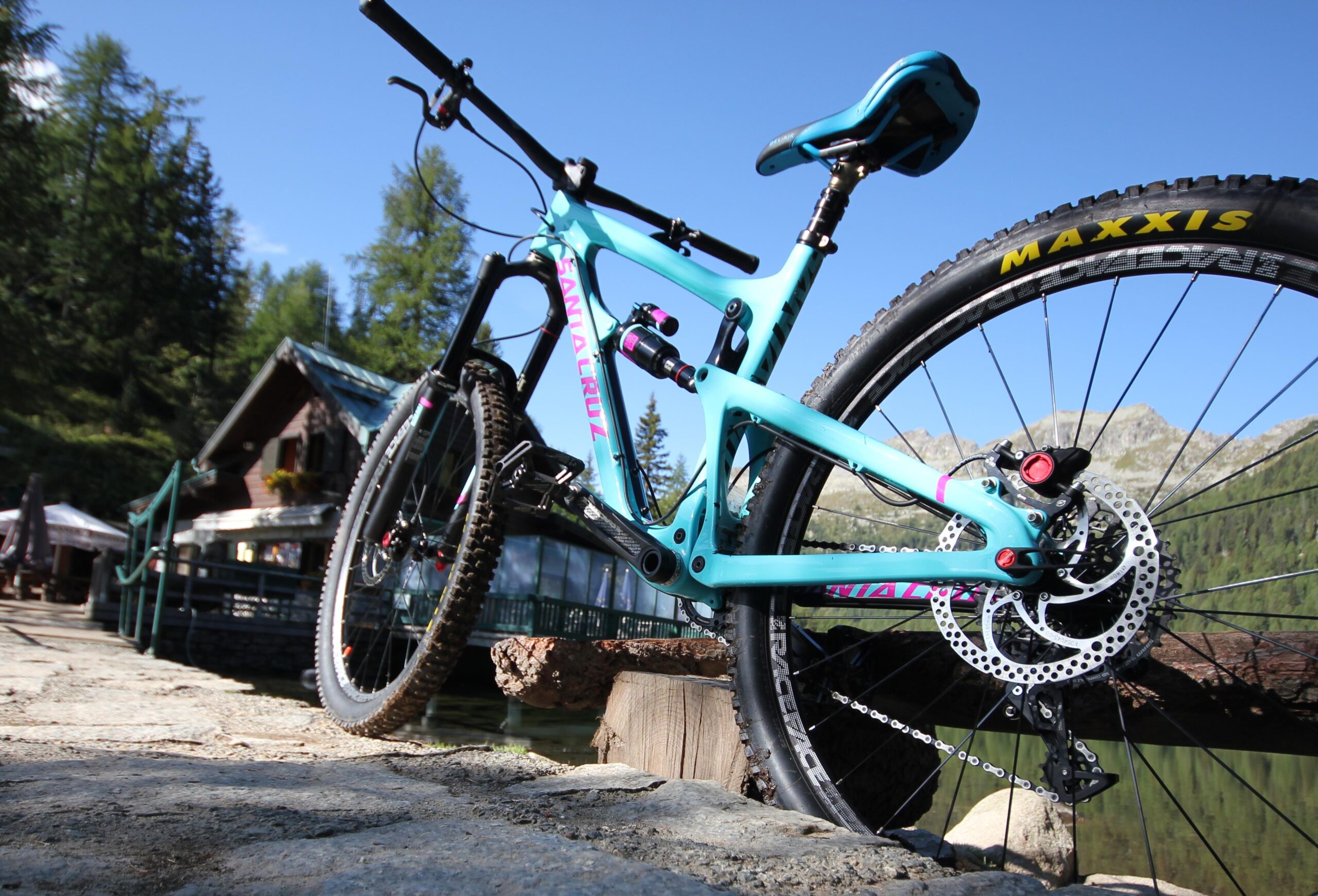 Santa Cruz Nomad: A mountain bike with a turquoise frame and pink accents resting on a rocky surface near a lake. In the background, a small wooden cabin is visible, surrounded by trees and mountains under a clear blue sky.
