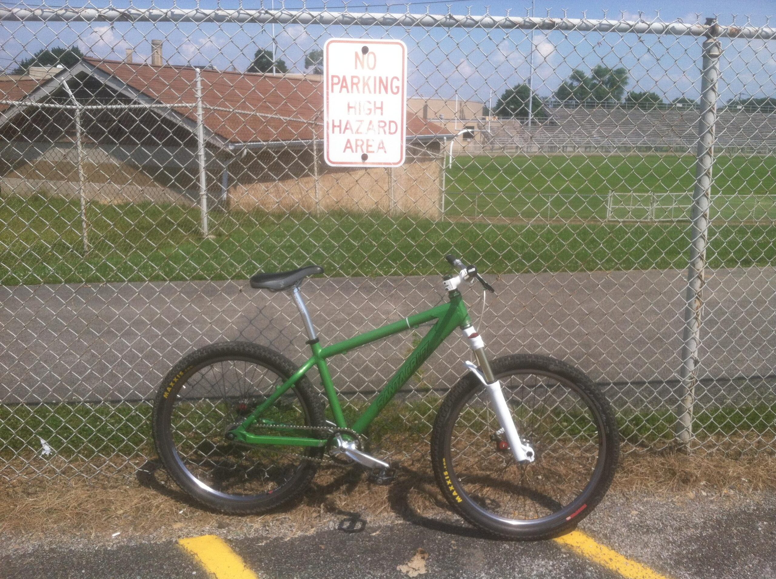 Santa Cruz Chameleon: A green mountain bike is leaning against a chain-link fence, with a "No Parking - High Hazard Area" sign visible above it. In the background, there is a grassy field and a building, with a few clouds in the blue sky. The ground is paved with yellow parking lines, though the bike is not parked in a designated spot.