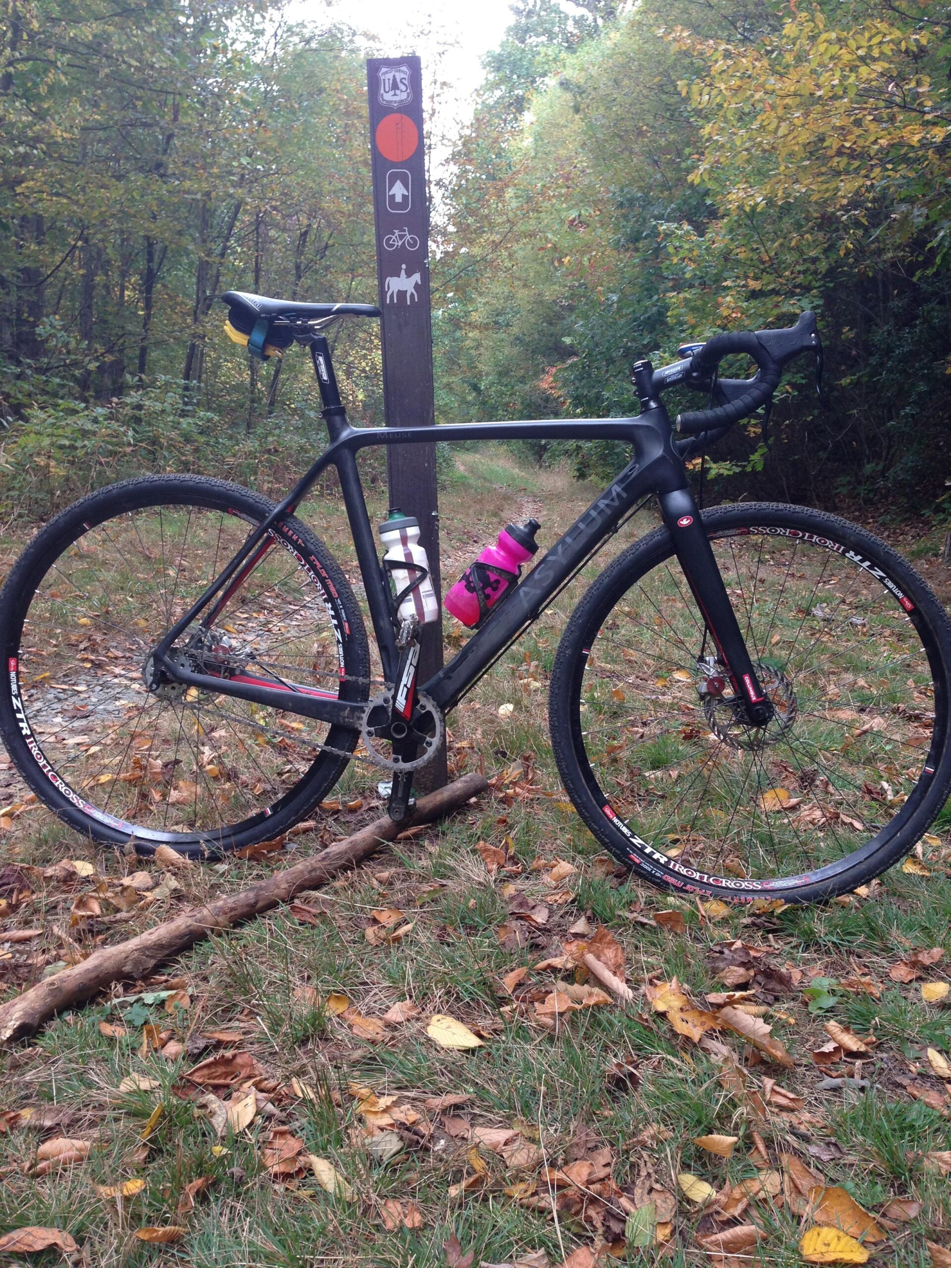 A black bicycle with red and white accents rests beside a trail sign in a wooded area. The sign indicates paths for hiking, biking, and horseback riding. Surrounding the bike and sign are autumn leaves scattered on the ground, with trees displaying various shades of green and yellow in the background. Clawhammer/5058 mountain bike trail.