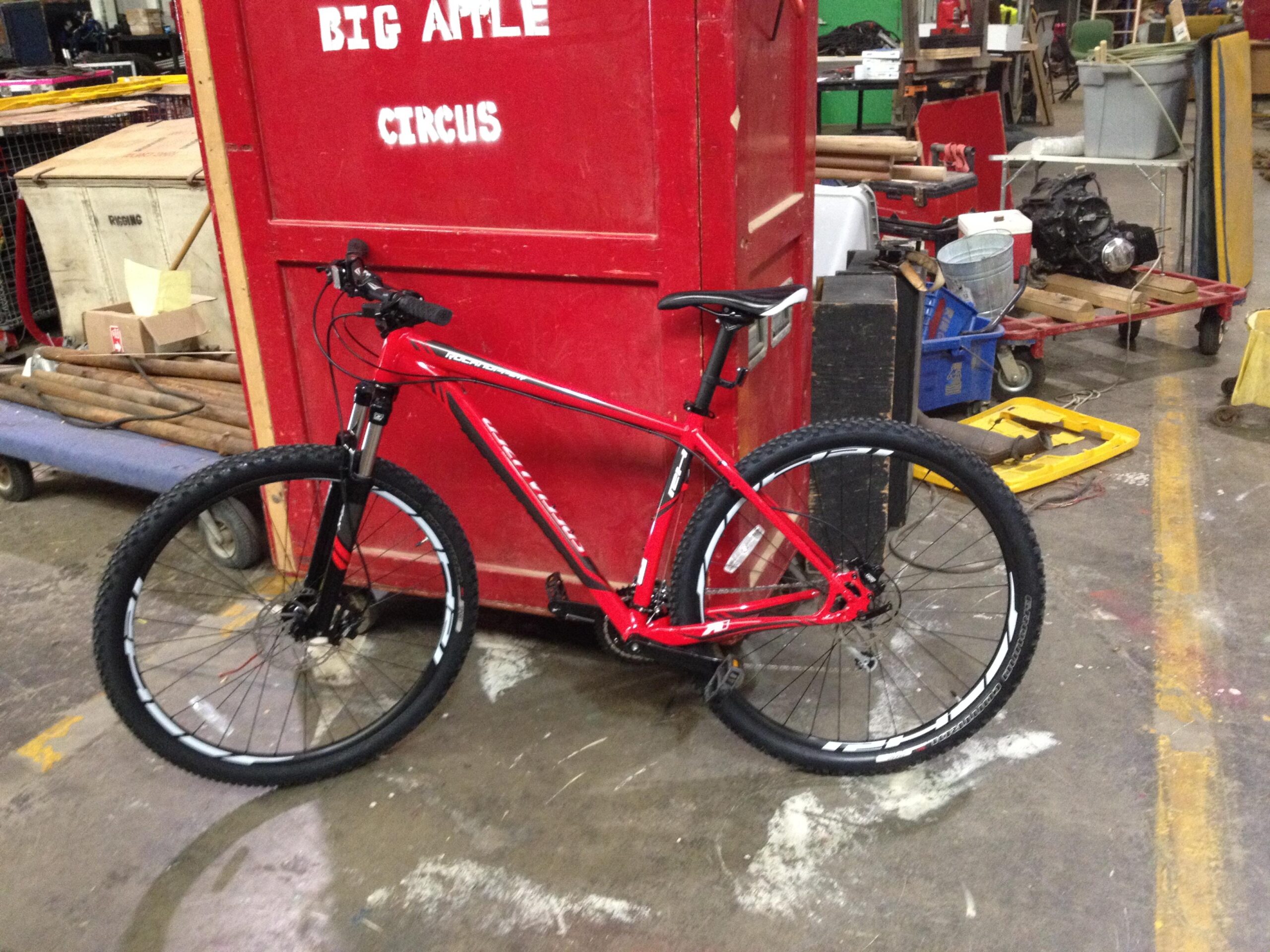 Specialized Rockhopper 29: A red mountain bike stands against a large red storage container in a cluttered workshop environment. The bike features thick tires and prominent front suspension, while various tools and equipment are scattered in the background.