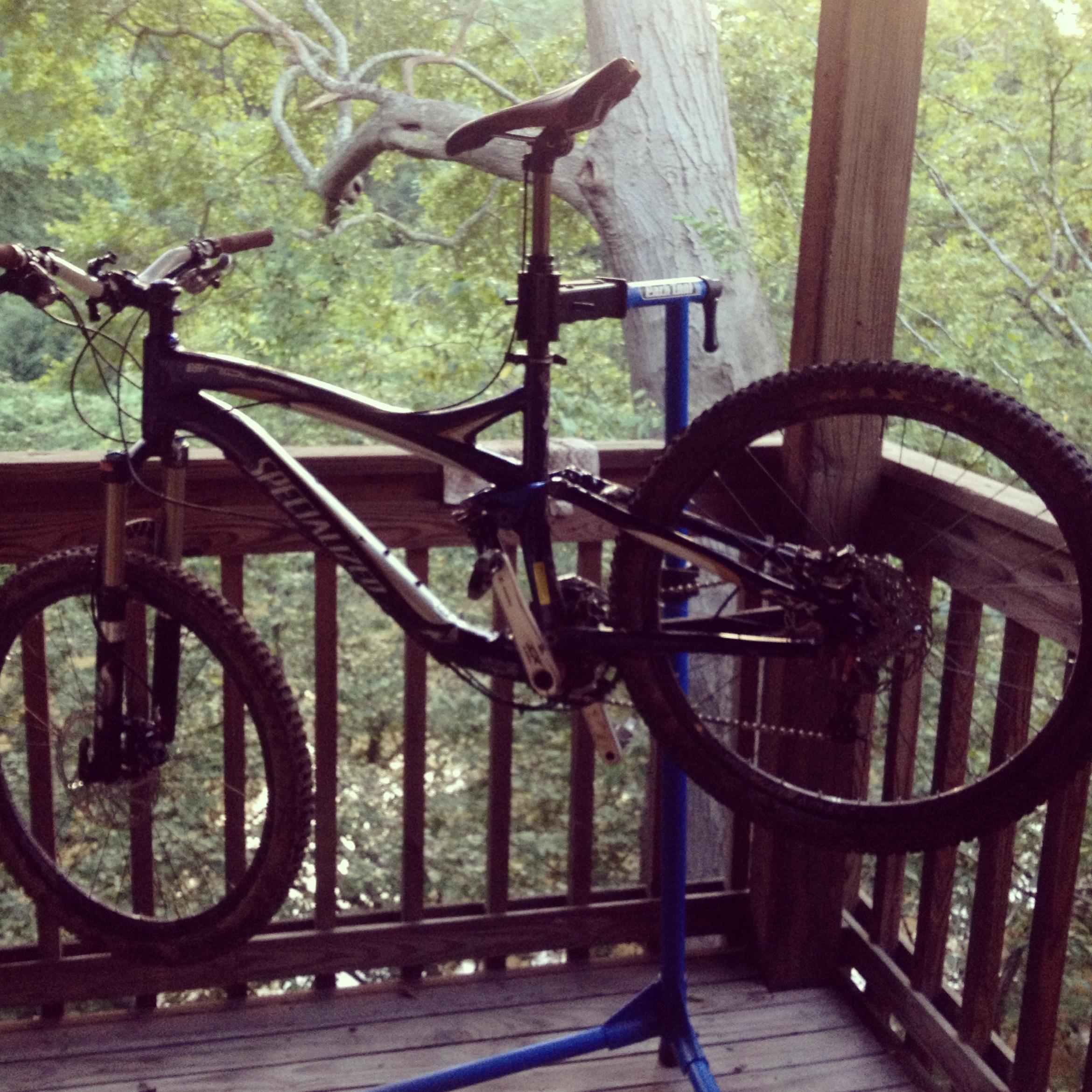 Specialized Enduro SL Pro: A mountain bike mounted on a repair stand on a wooden balcony, with trees visible in the background. The bike features a black and blue frame and is positioned with its rear wheel off the ground, suggesting maintenance is in progress.