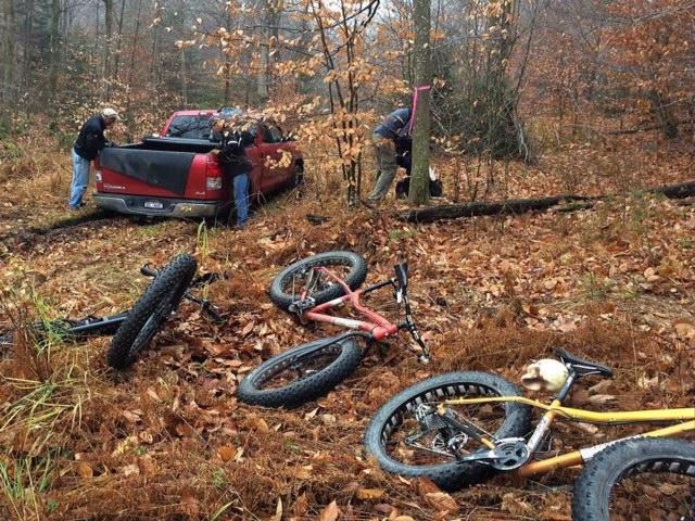 A group of people is near a red pickup truck parked in a forested area during autumn. Two individuals are working near the truck, while a third is unloading items from the truck bed. Several bicycles are scattered on the ground, surrounded by fallen leaves. Trees with bare branches can be seen in the background.