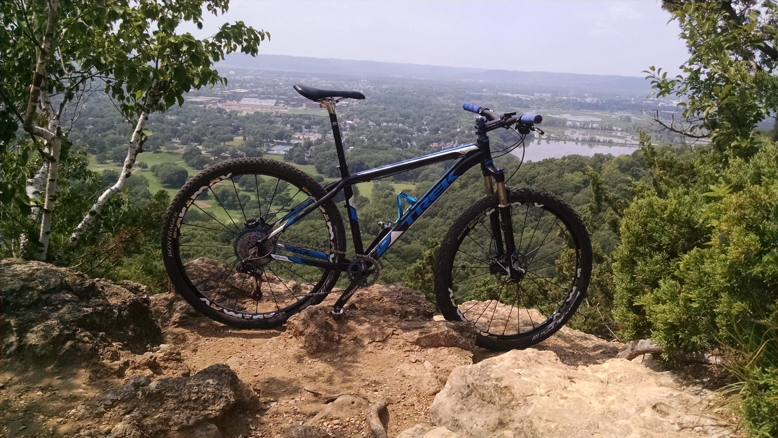 Trek X-Caliber: A mountain bike resting on rocks overlooking a lush green valley and distant hills under a clear sky. The bike has a black and blue frame and is positioned prominently in the foreground, with scenic views of trees and a river in the background.