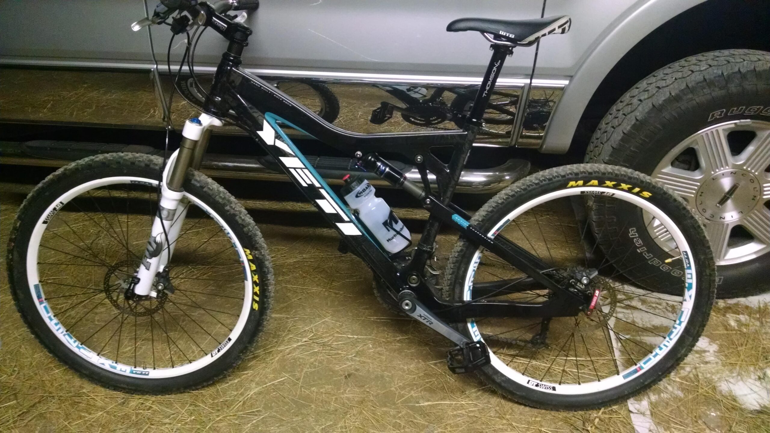 Yeti ASR 5 Carbon: A black mountain bike with white and blue accents is positioned on a bed of straw. The bike features thick tires with "Maxxis" branding, a water bottle mounted on the frame, and a visible suspension fork. A silver truck is partially visible in the background, suggesting an outdoor setting.