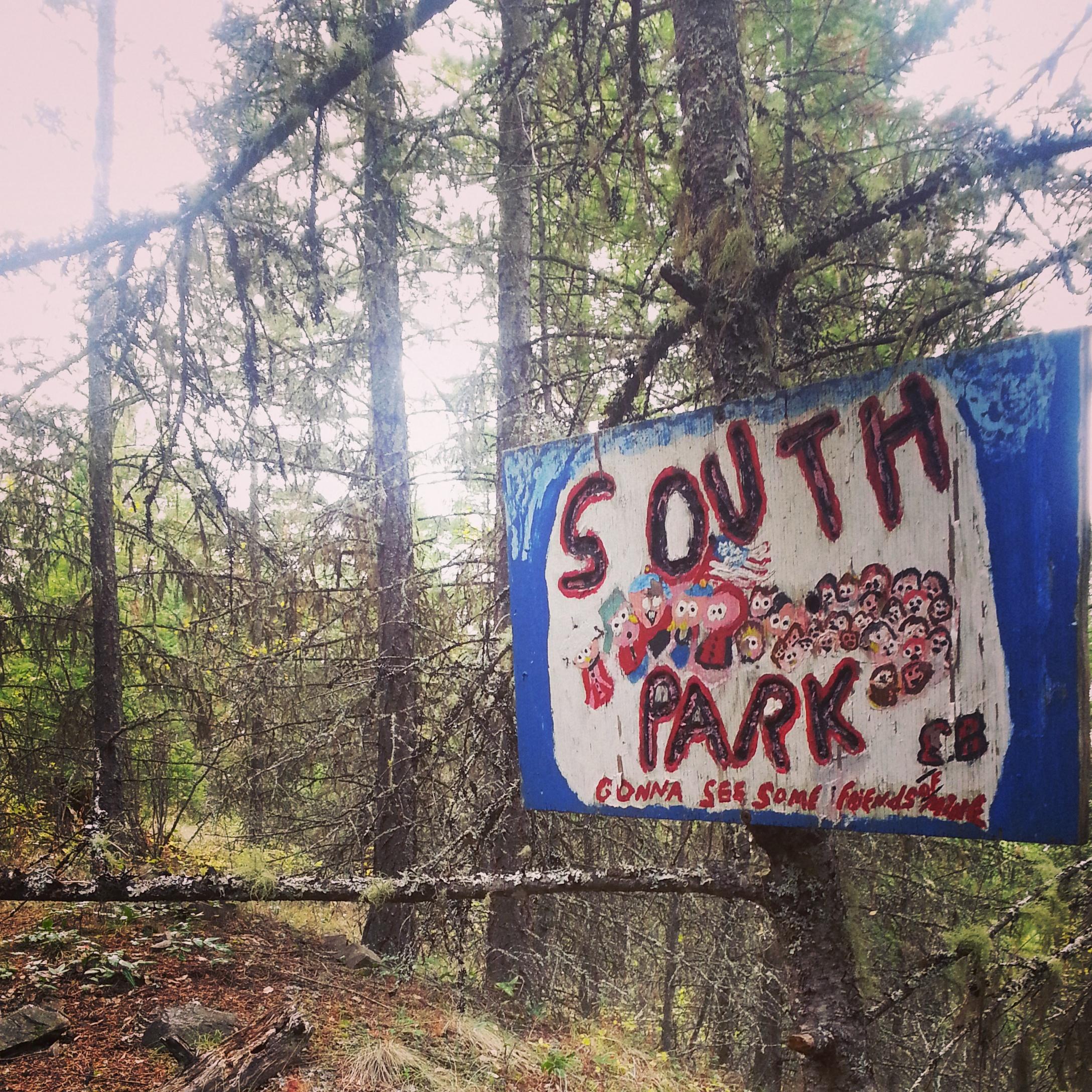 A hand-painted wooden sign that reads "SOUTH PARK" with colorful, whimsical characters depicted. The sign is located in a densely wooded area, surrounded by tall, green trees. The atmosphere appears slightly overcast, suggesting a peaceful, natural setting. The lower portion of the sign includes the phrase "gonna see some friends." Kalamalka Lake Provincial Park mountain bike trail.