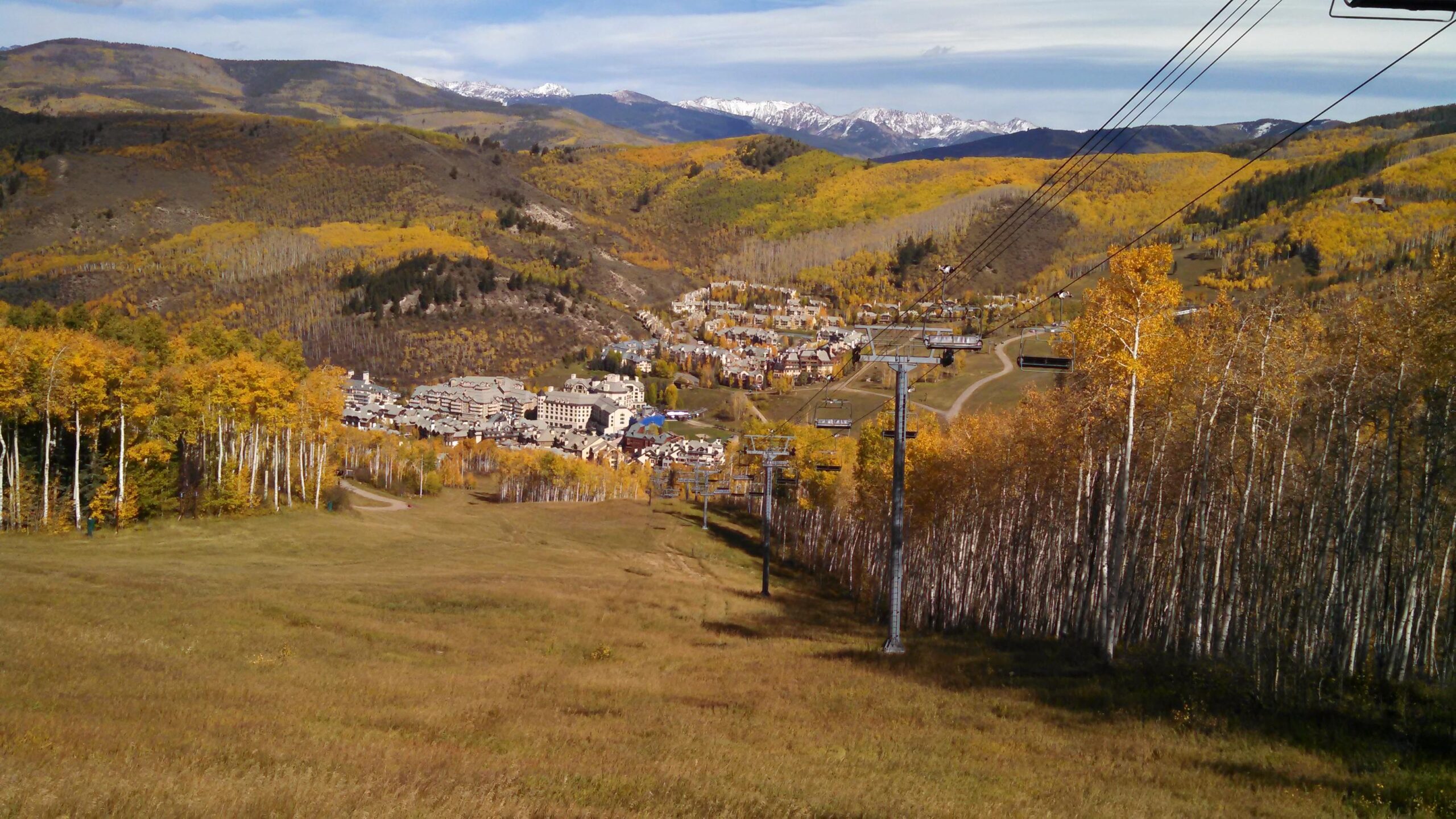 A picturesque mountain landscape featuring vibrant autumn foliage, with golden aspen trees in the foreground and a view of a ski resort nestled in the valley below. Snow-capped mountains are visible in the distance under a partly cloudy sky. Ski lift chairs are seen along the hillside, indicating a popular winter sports destination. Beaver Creek Ski Resort mountain bike trail.