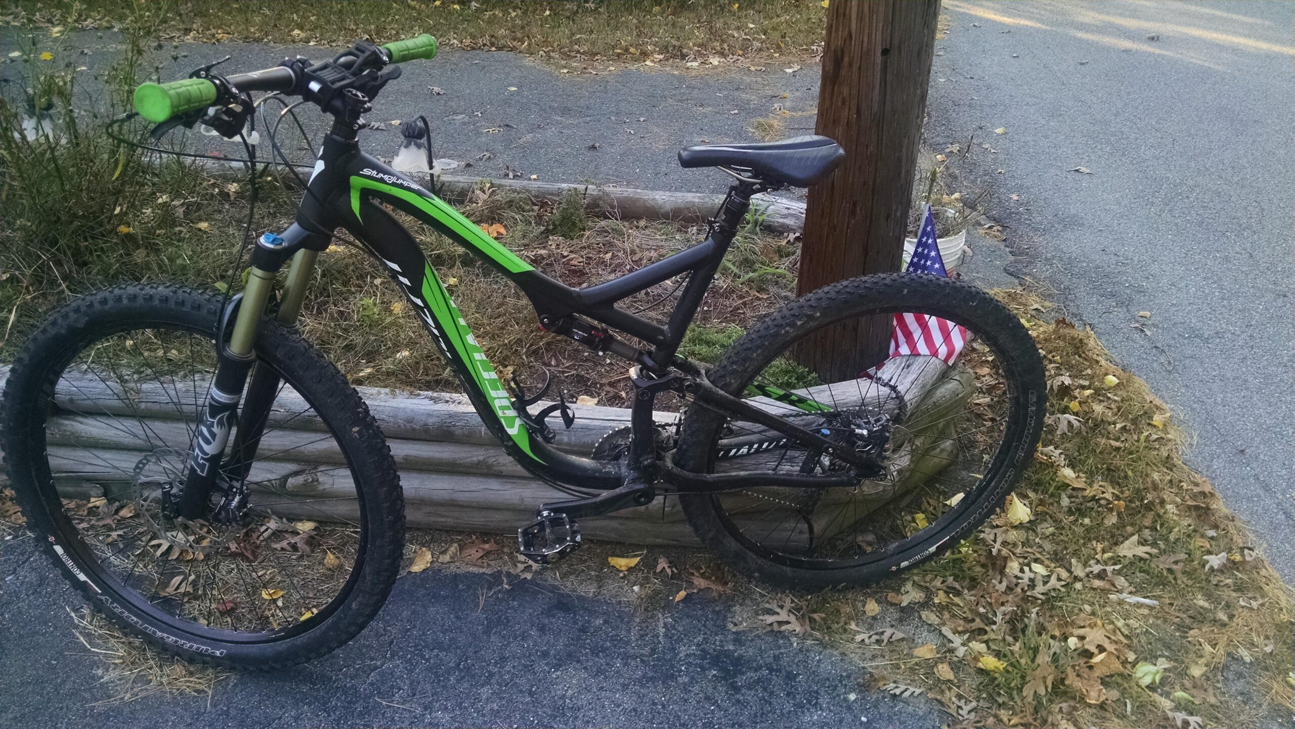 Specialized Stumpjumper: A black and green mountain bike parked next to a wooden post, with a small American flag visible in the background. The bike features green grips, a sturdy frame, and thick tires, resting on a paved path surrounded by autumn leaves.
