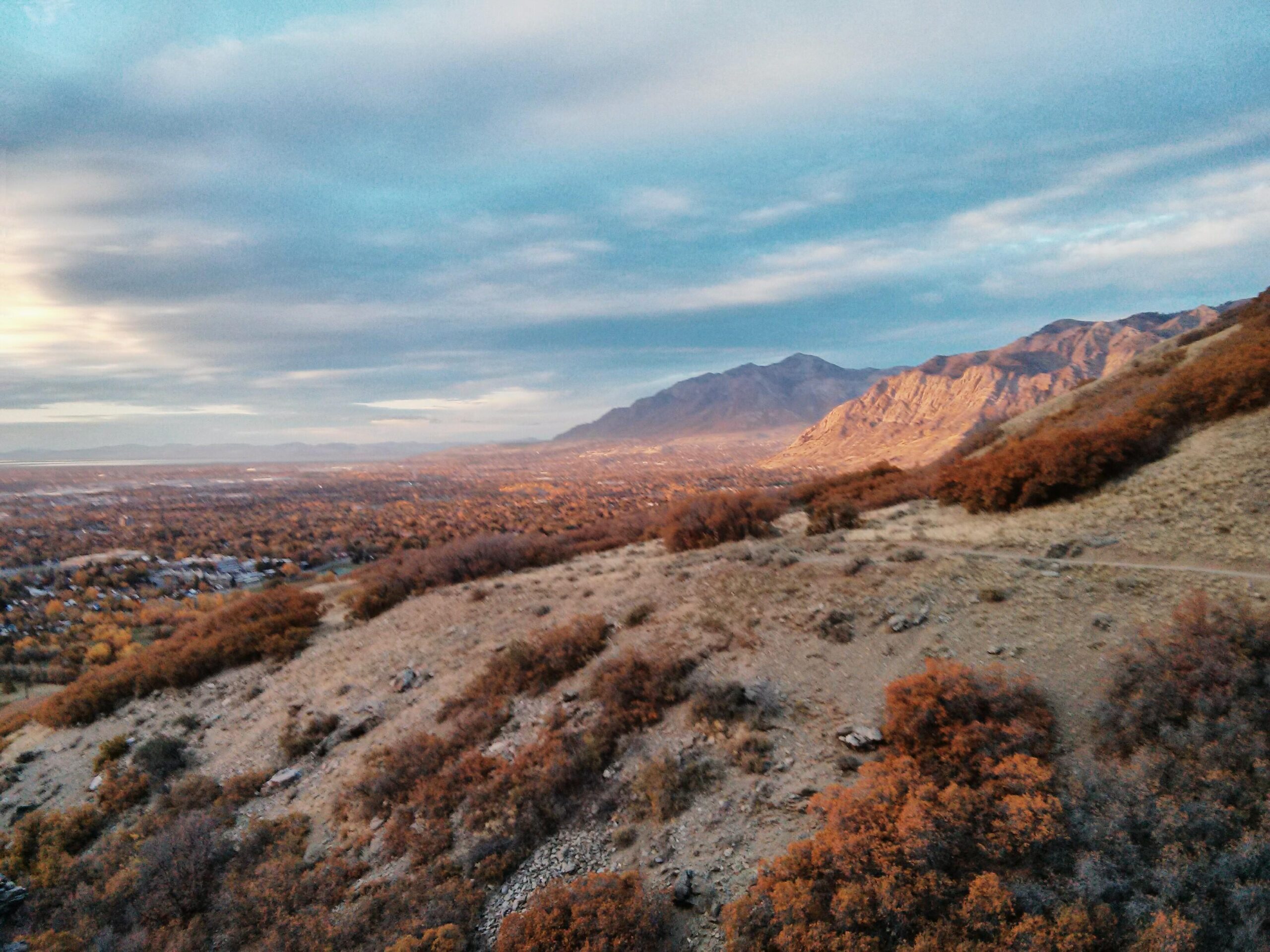 A scenic view of a valley surrounded by mountains, showcasing rolling hills with autumn foliage in shades of orange and yellow. The sky is partly cloudy with soft blue hues, and a small town can be seen nestled in the valley below. The landscape captures the tranquil beauty of nature during the fall season. Bonneville Shoreline Ogden South Of 12th mountain bike trail.