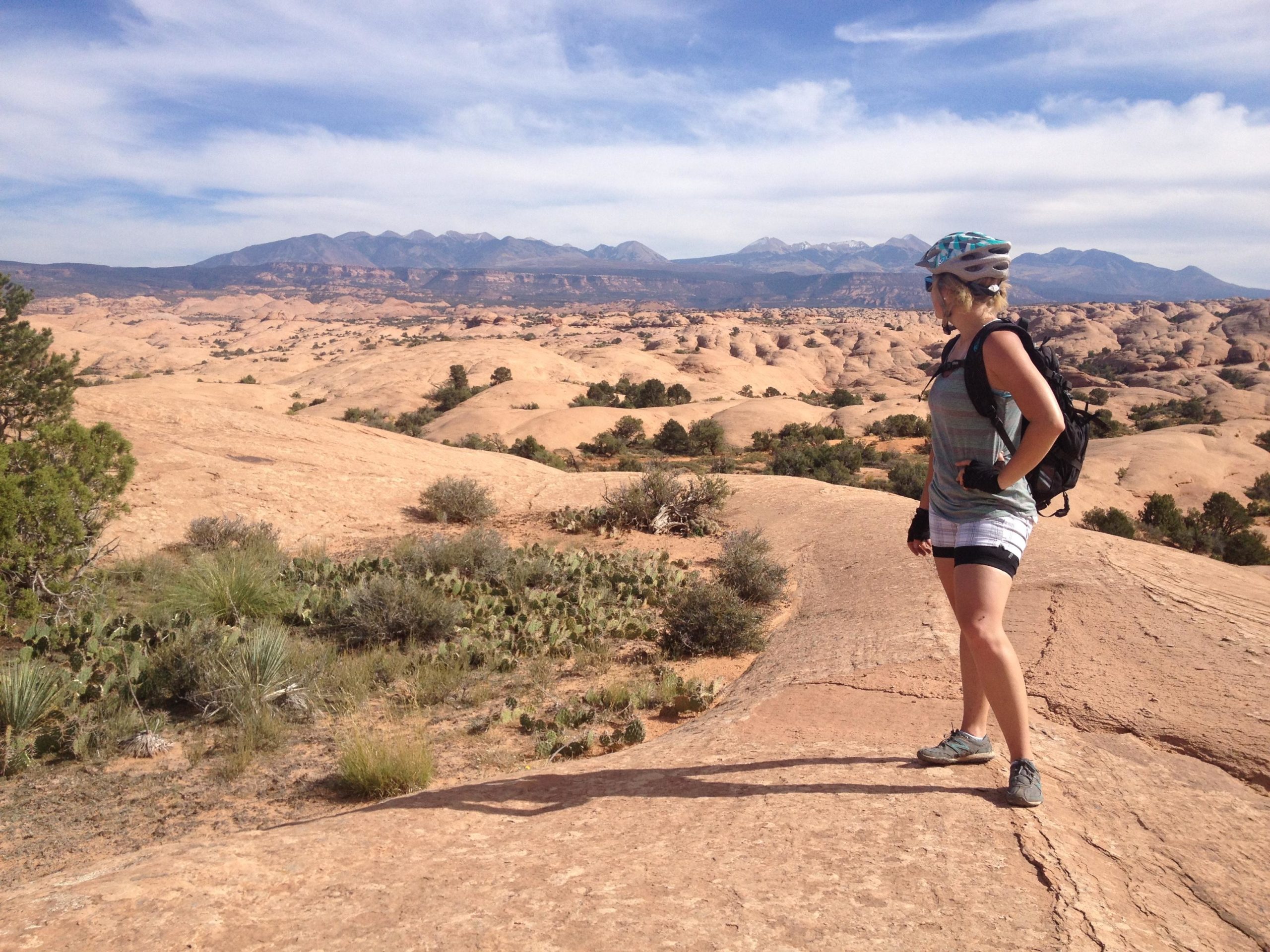 A person in a cycling helmet stands on rocky terrain, overlooking a vast landscape of rolling hills and distant mountains. The scenery features sparse vegetation, including cacti and shrubs, under a blue sky with a few clouds. Slickrock mountain bike trail.