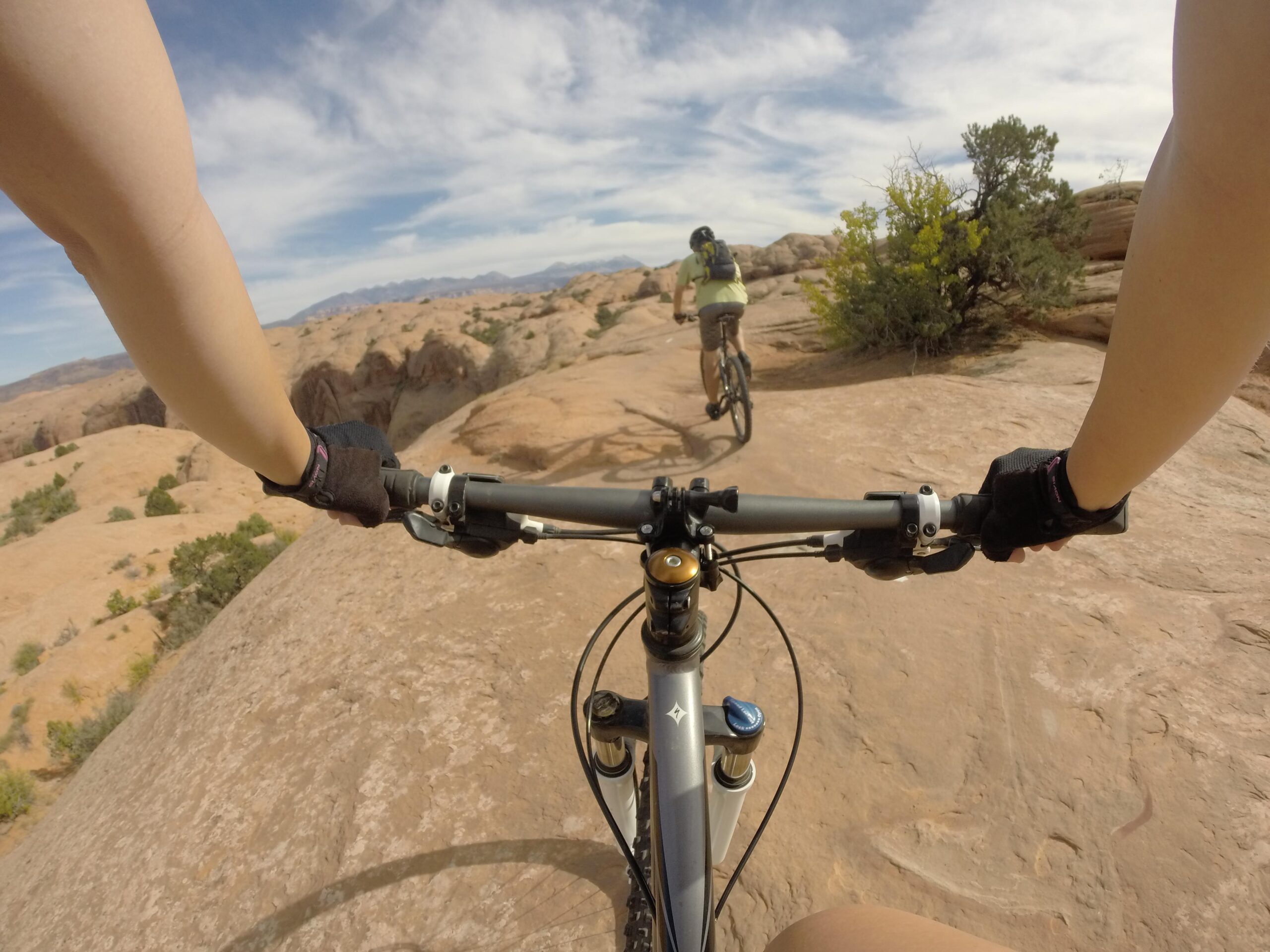 A first-person view of a mountain biking trail, capturing the hands gripping the handlebars of a bike with a rocky landscape in the background. A second cyclist is visible in the distance, navigating the rugged terrain under a blue sky with wispy clouds. Slickrock mountain bike trail.