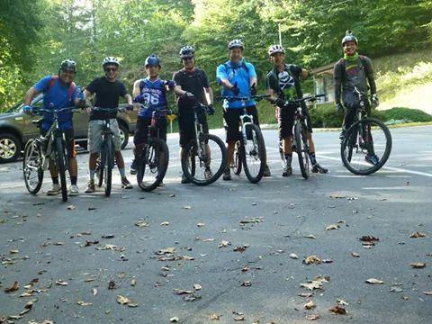 A group of six cyclists pose for a photo in a parking area, each wearing biking gear and standing next to their mountain bikes. The background features trees and a sunny, green landscape, with fallen leaves scattered on the ground. Warrior Creek mountain bike trail.