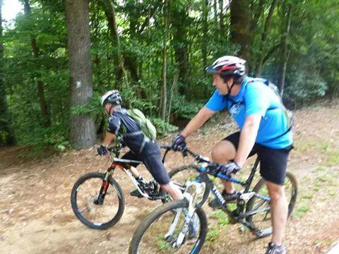 Two cyclists navigating a wooded trail. One cyclist, wearing a backpack and helmet, is leaning forward on their bike, while the other, dressed in a blue shirt, is positioned on a mountain bike, looking ahead. Tall trees and greenery surround the trail, suggesting a natural, outdoor environment. Warrior Creek mountain bike trail.