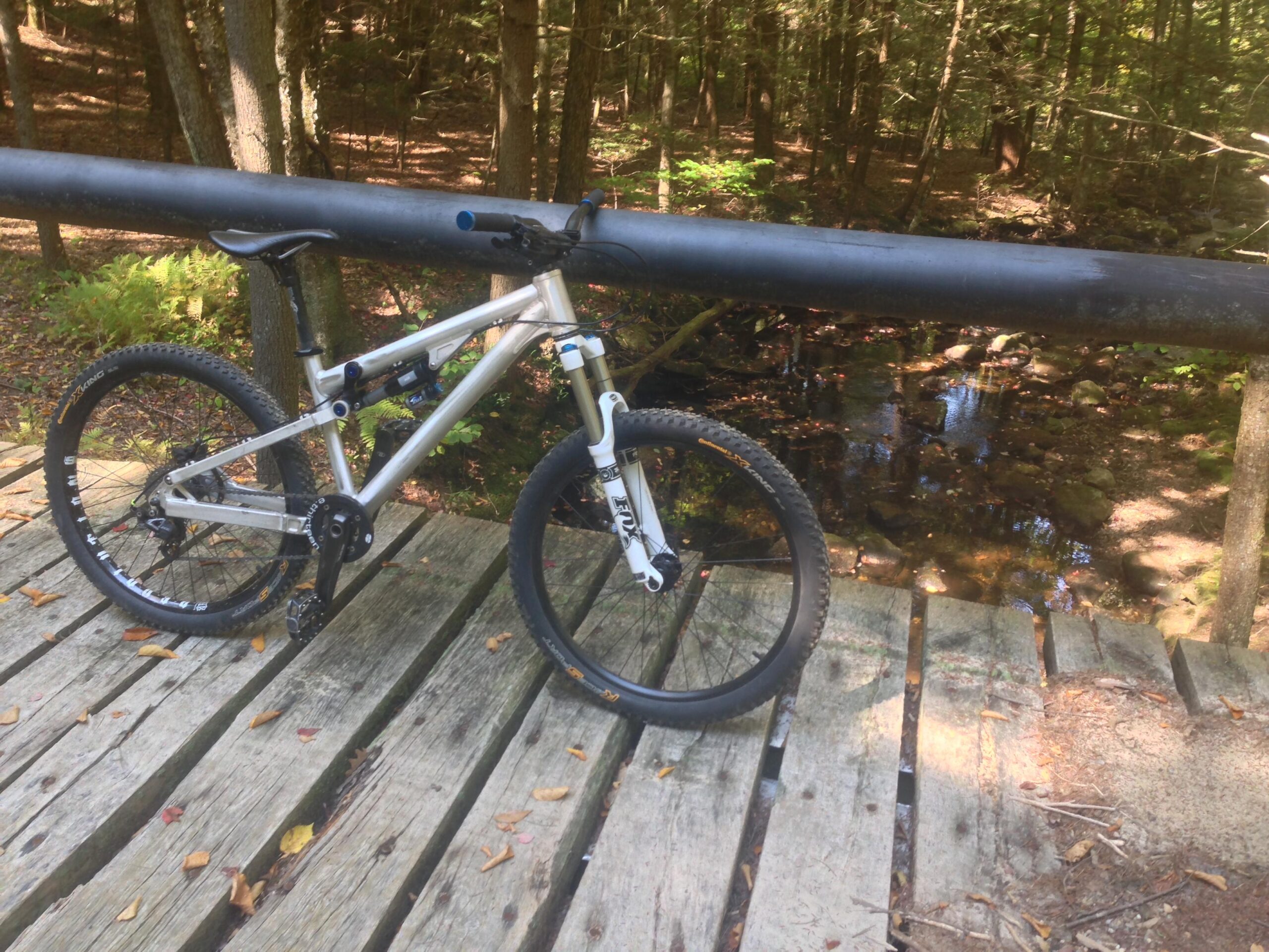 Transition Bottlerocket: A silver mountain bike parked on a wooden bridge overlooking a small stream, surrounded by lush green trees and foliage. The scene captures the tranquility of a wooded area perfect for outdoor activities.