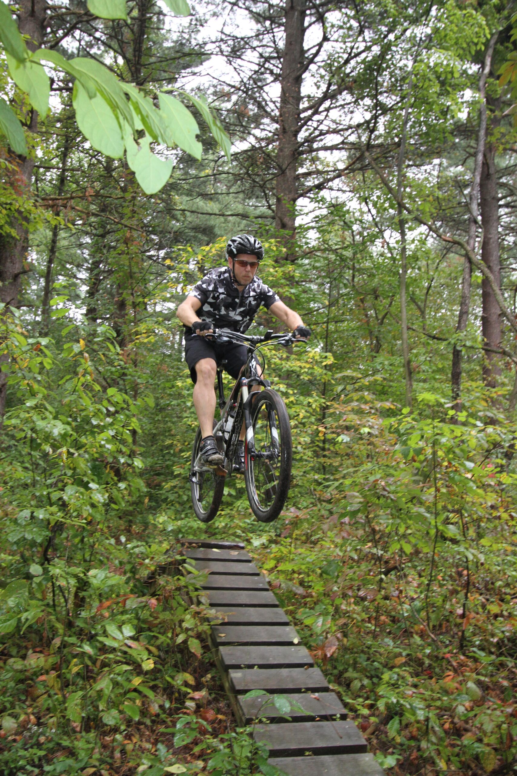 A mountain biker in a camouflage shirt and helmet performs a jump over a wooden plank in a wooded area, surrounded by lush greenery and trees. The scene captures the excitement of off-road cycling in a natural setting. Van  Buren mountain bike trail.