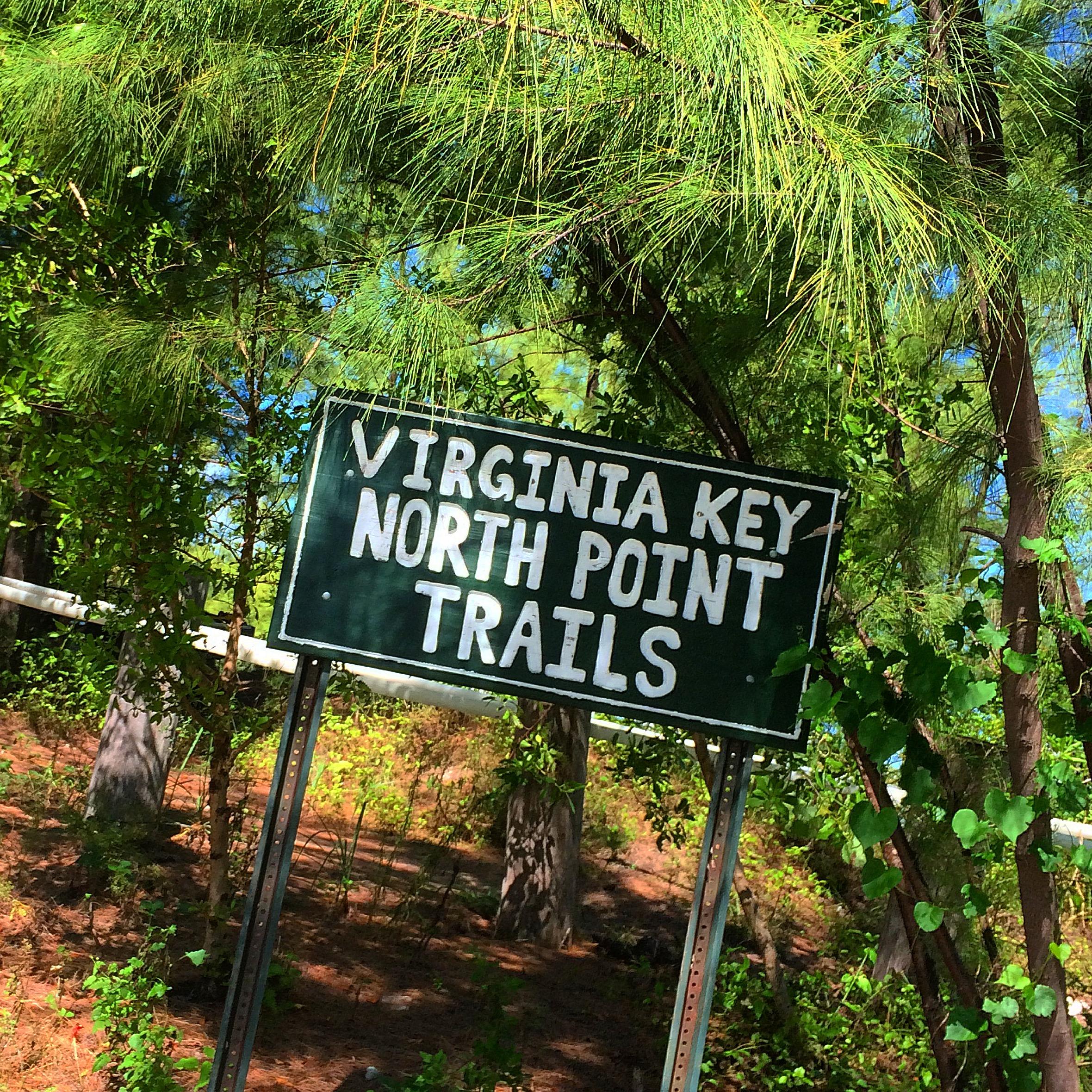 A green sign with white text reading "Virginia Key North Point Trails" is surrounded by lush greenery and trees, indicating the location of walking or hiking trails. Virginia Key North Point mountain bike trail.
