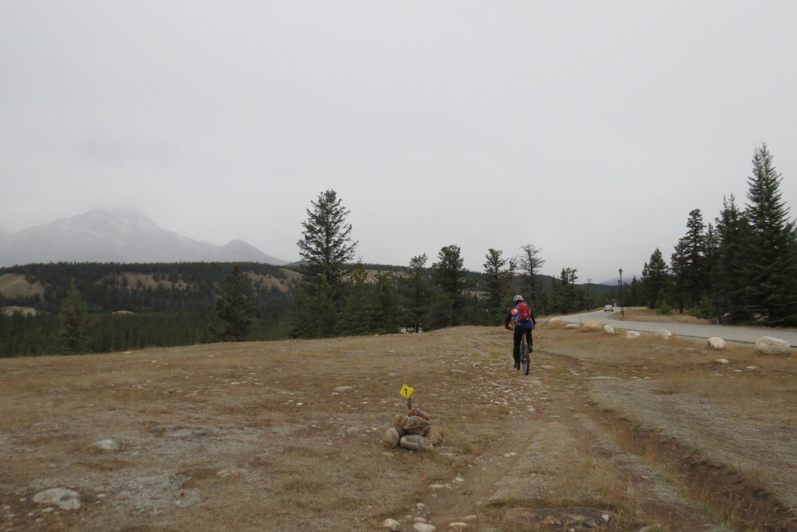 A person riding a bicycle along a dirt path in a scenic mountainous area, surrounded by evergreen trees and rocky terrain. The sky is overcast, and distant mountains are visible in the background. A trail marker with the number "1" is positioned on a small pile of rocks next to the path. A paved road can be seen in the distance. Trail 7 / 7H (Teahouse) mountain bike trail.