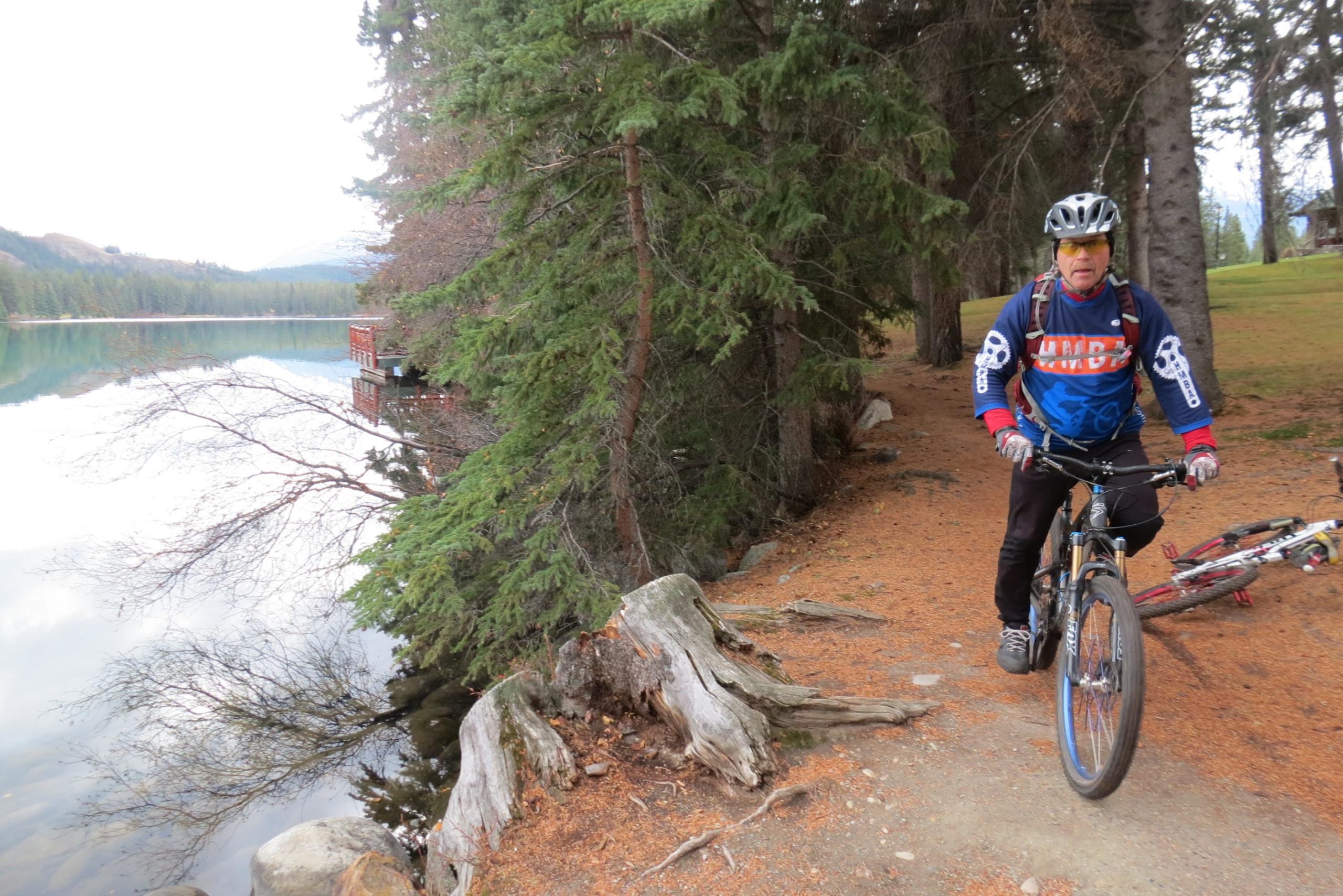 A cyclist wearing a helmet and a colorful long-sleeve shirt stands beside their mountain bike on a path near a tranquil lake surrounded by pine trees. The calm water reflects the forest and distant mountains, creating a serene outdoor scene. Trail 7 / 7H (Teahouse) mountain bike trail.