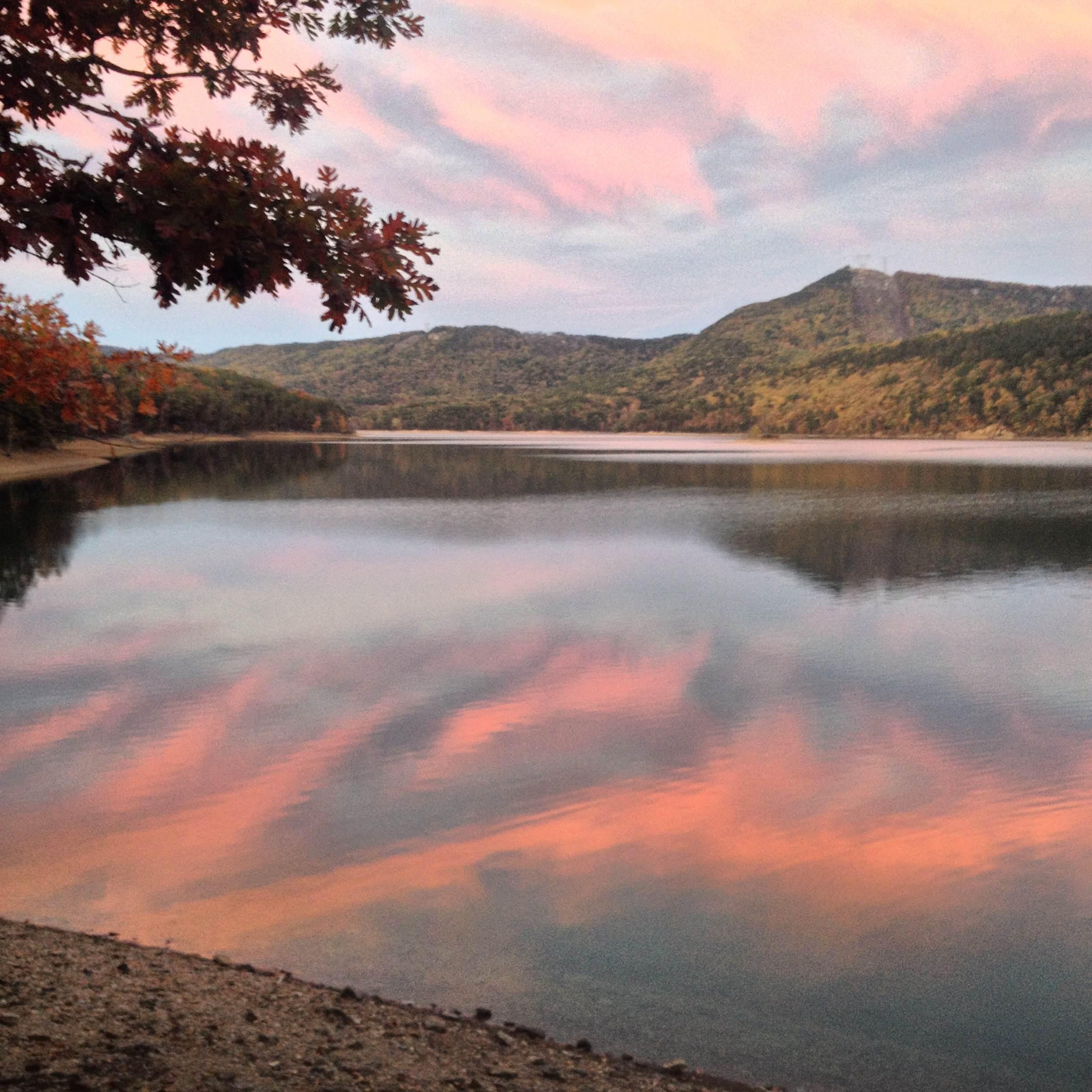 A tranquil lakeside scene at sunset, featuring still water reflecting soft pink and blue clouds against a backdrop of rolling hills. Bright autumn foliage lines the water's edge, enhancing the serene atmosphere. Lakeside Trail mountain bike trail.