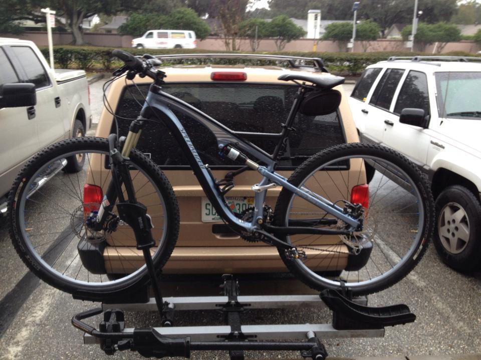 Specialized Camber 29: Alt text: A mountain bike is securely mounted on a bike rack attached to the rear of a tan SUV in a parking lot. Other vehicles, including a gray truck and a white SUV, are visible in the background. The scene is slightly rainy, with water droplets on the bike.