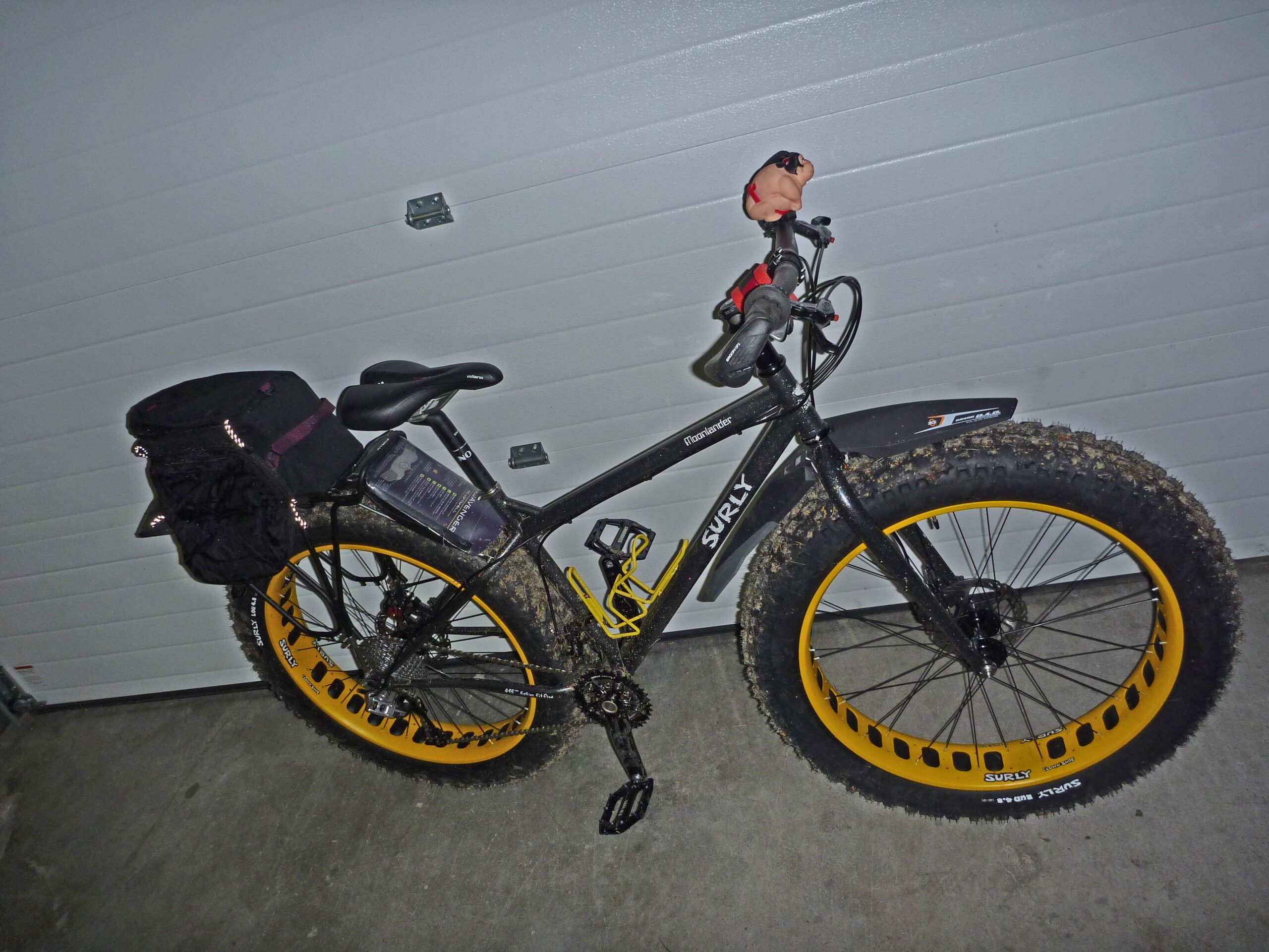 Surly Moonlander: A black Surly bike with oversized yellow rims is parked against a garage wall. The bike features a black bag on the rear rack and has muddy tires, indicating recent outdoor use. A small figure is attached to the handlebars.