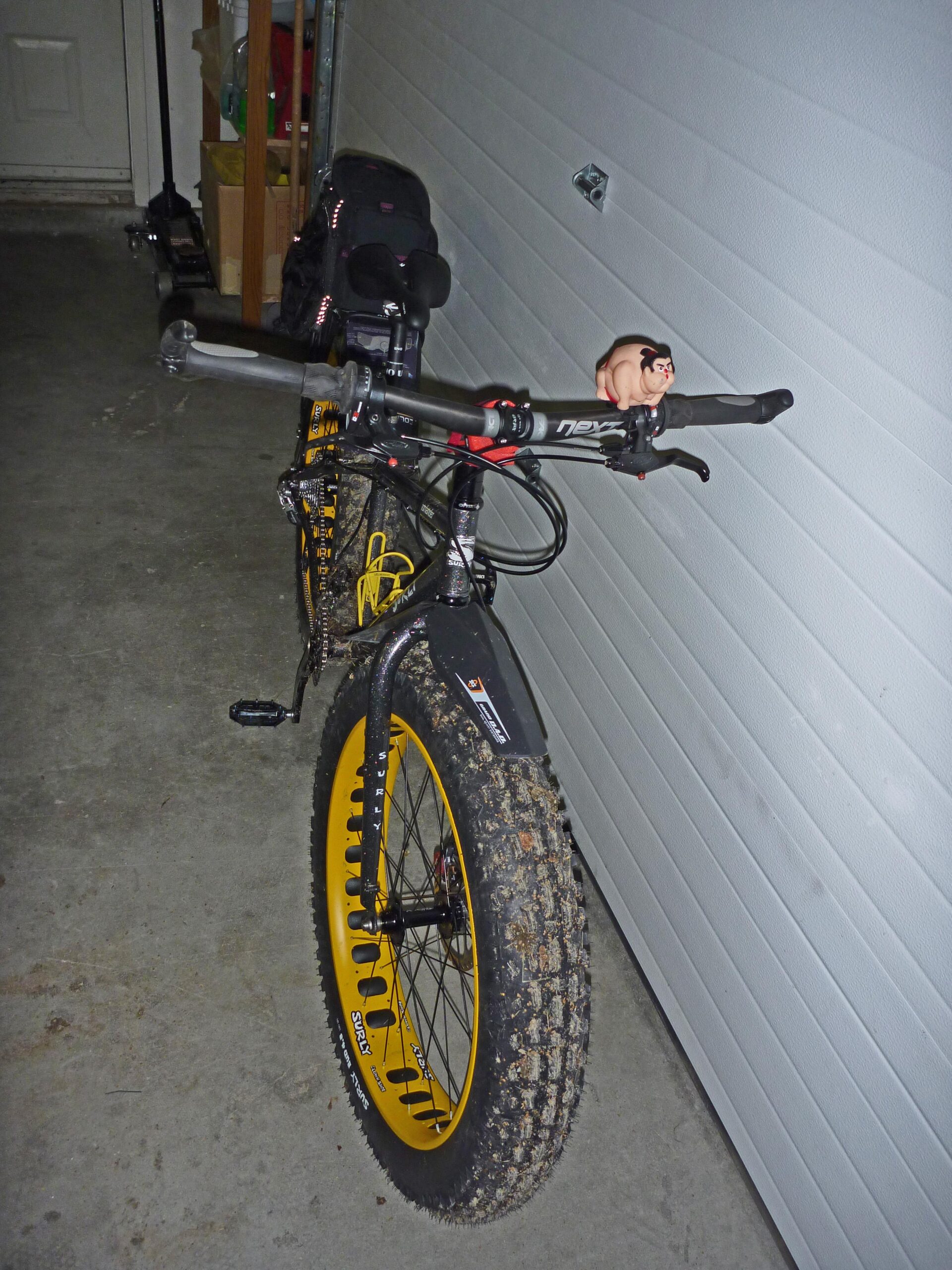 Surly Moonlander: A fat bike parked in a garage, featuring wide tires with distinctive yellow rims. It has a black seat and handlebars adorned with a small, humorous figurine. The background includes a closed garage door and some storage items.