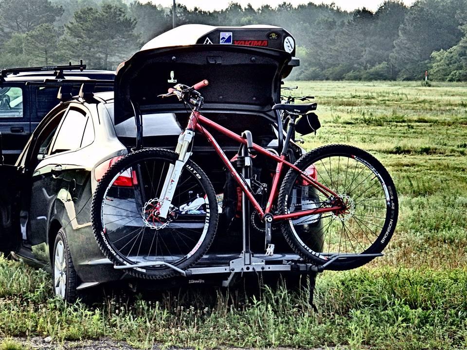 Niner S.I.R. 9: A red mountain bike mounted on a bike rack attached to the back of a black car with the trunk open. The background features a grassy area and distant trees, suggesting an outdoor setting.