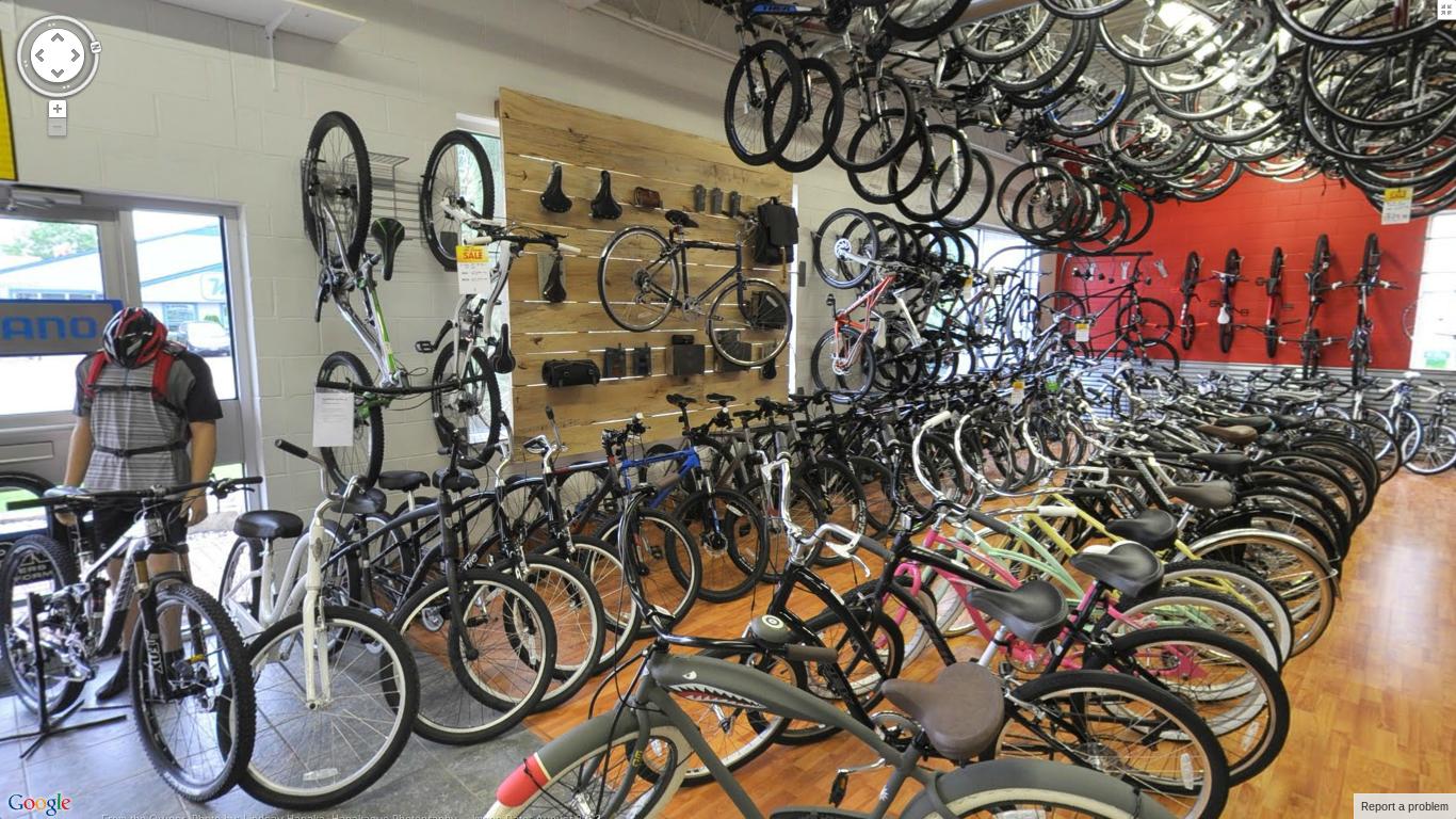 A busy bicycle shop interior featuring a wide variety of bicycles on display. The shop has bikes arranged on the floor and hanging on walls, with a focus on both mountain and road bikes. A customer is seen browsing among the bikes, while a wooden display board showcases bike accessories. The space is bright and inviting, with a warm wooden floor and colorful bicycles in various styles.