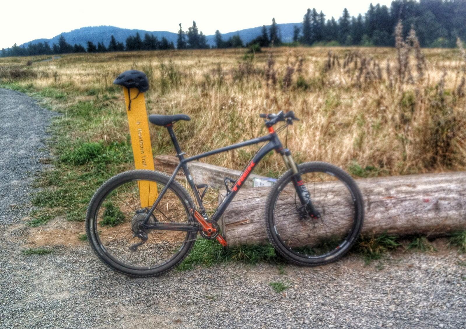 A mountain bike leaning against a log on a gravel trail, with a yellow trail sign and a black helmet resting on top of the sign. In the background, a grassy field and distant mountains are visible under an overcast sky. Powell Butte Nature Park mountain bike trail.