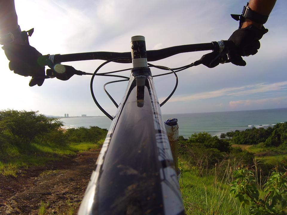 A close-up view from the handlebars of a mountain bike, overlooking a coastal landscape with the ocean in the background and a clear sky. The hands gripping the handlebars are gloved, and the scene captures a sense of adventure and nature. Corredor Ecologico mountain bike trail.