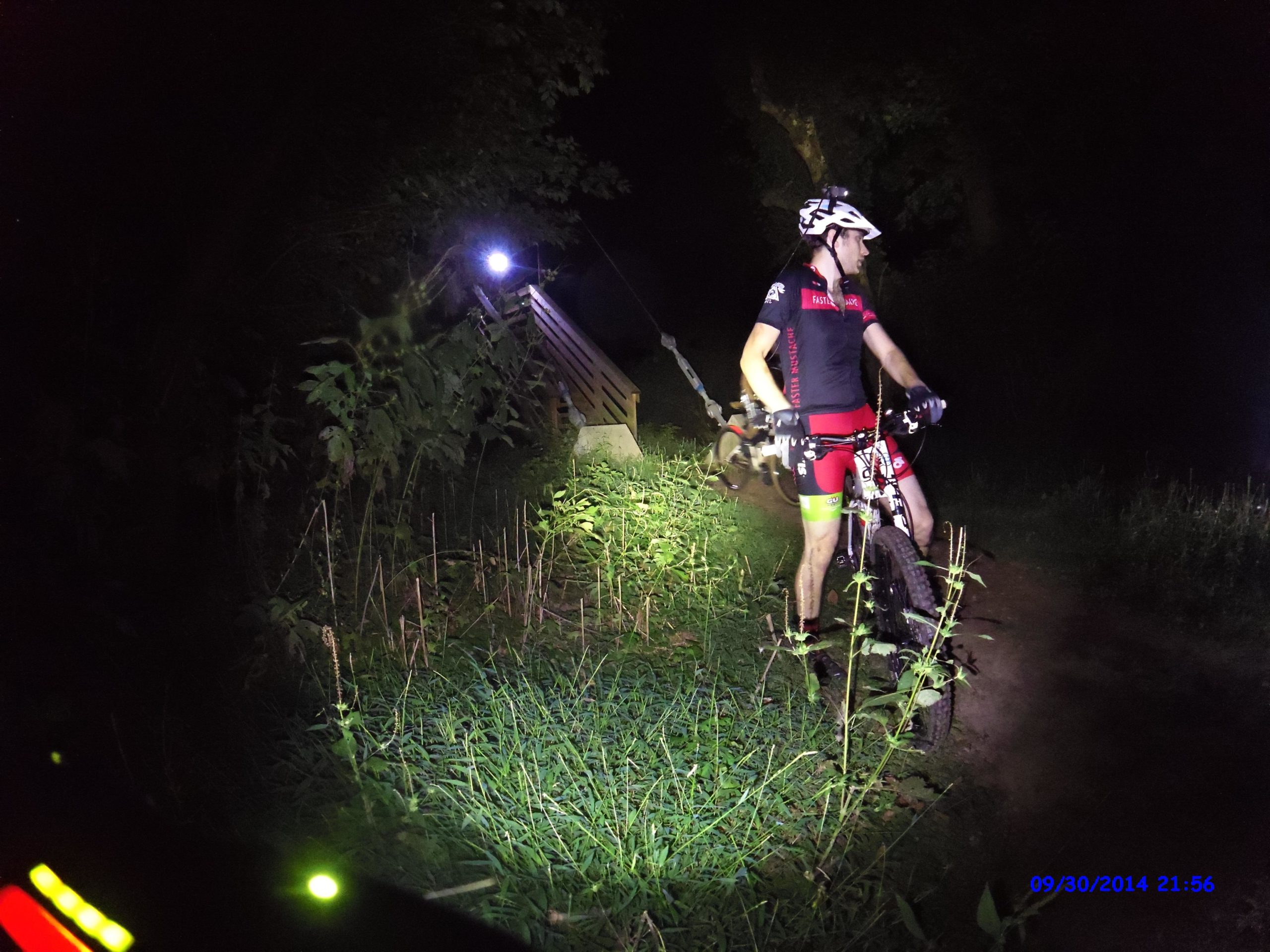 A mountain biker in a red and black outfit stands on a narrow, grassy trail illuminated by bike lights at night. The rider is facing away from the camera, and a wooden structure is visible in the background. The scene is surrounded by trees and tall grass, creating a nighttime outdoor atmosphere. Morningside Nature Preserve mountain bike trail.