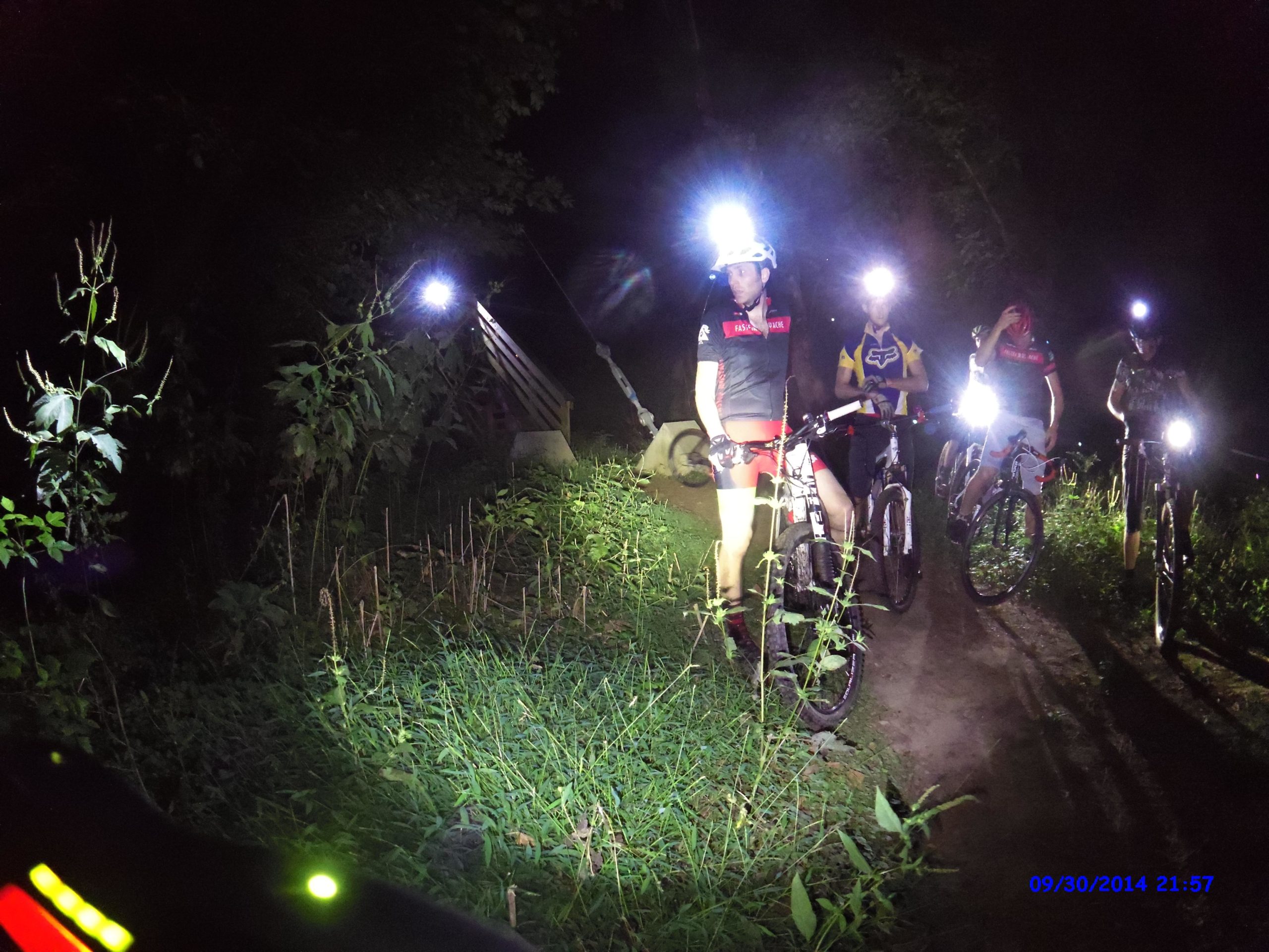 A group of mountain bikers dressed in athletic gear are gathered on a dirt path at night, illuminated by headlamps. Surrounding vegetation and grass are partially lit, creating a contrast with the darkness of the surrounding area. The scene conveys a sense of adventure and camaraderie in an outdoor nighttime setting. Morningside Nature Preserve mountain bike trail.
