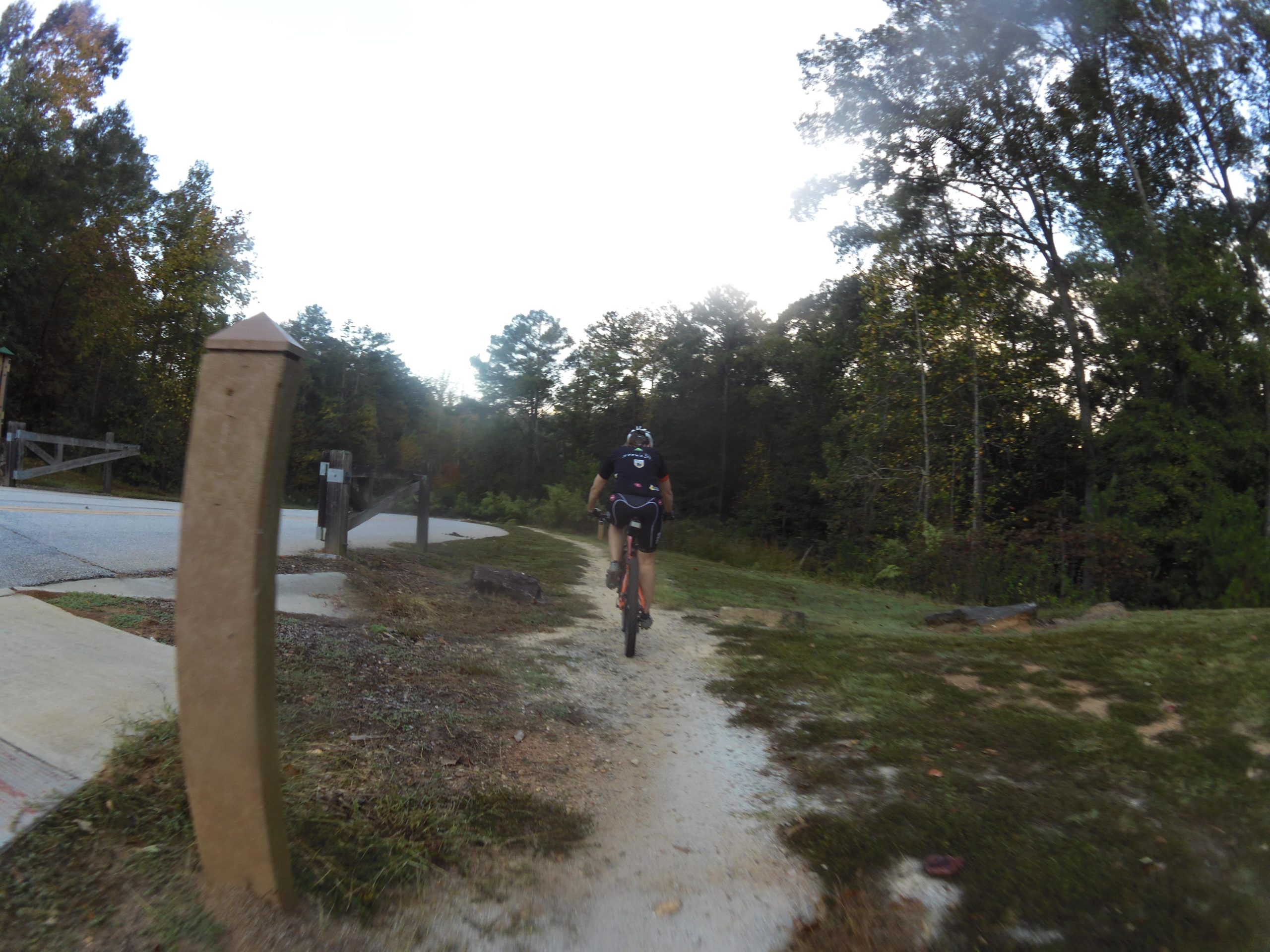 A person riding a mountain bike on a gravel path surrounded by trees, with a wooden post in the foreground and a park entrance in the background. The scene is set in a natural environment during the early morning or late afternoon. Yellow River mountain bike trail.