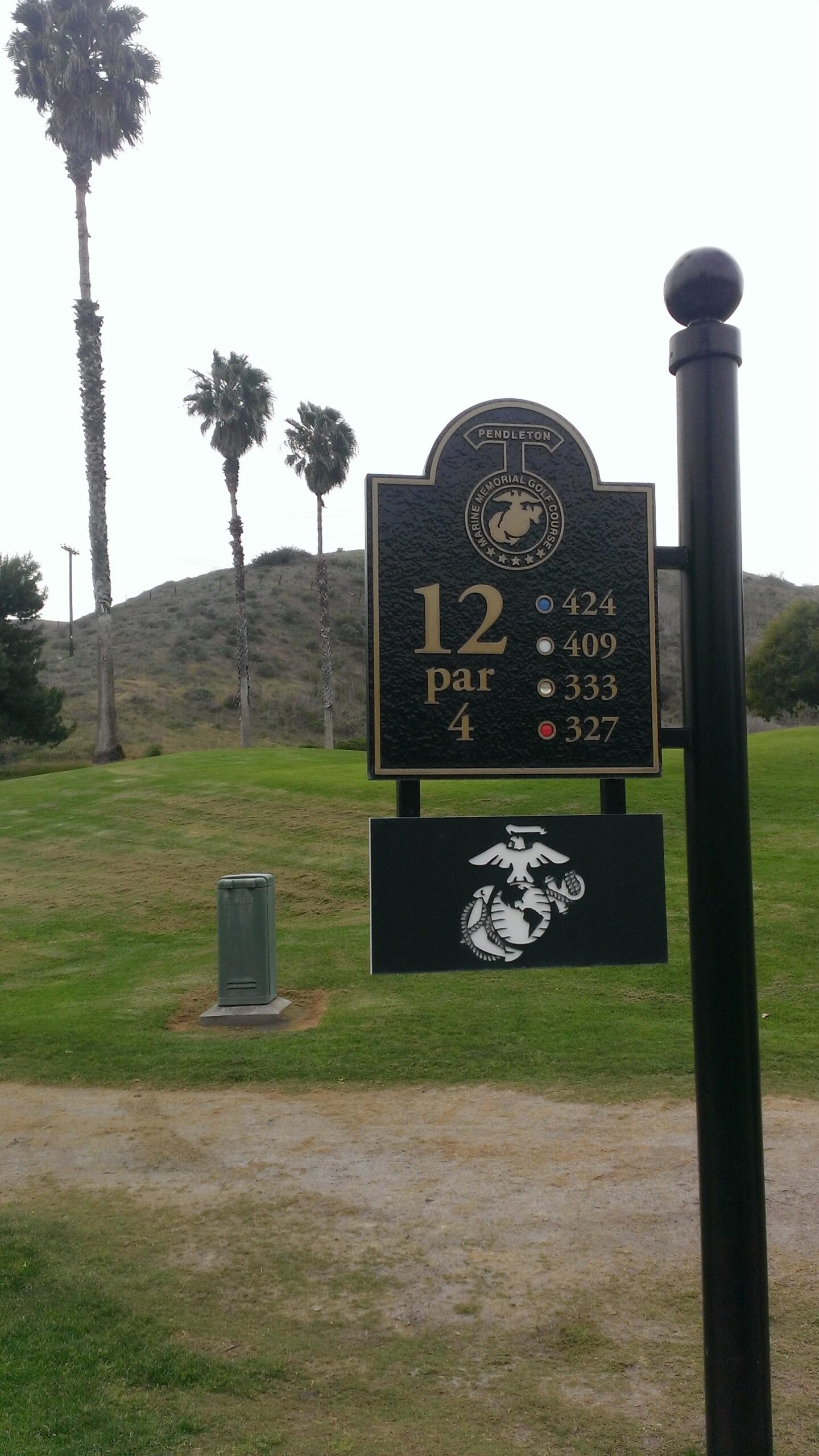A sign at Pendleton Marine Memorial Golf Course indicating the 12th hole, a par 4. The sign displays distance markings: 424 yards (blue tee), 409 yards (white tee), 333 yards (gold tee), and 327 yards (red tee). In the background, palm trees are visible, and there is a grassy area. MC Memorial Golf Course mountain bike trail.