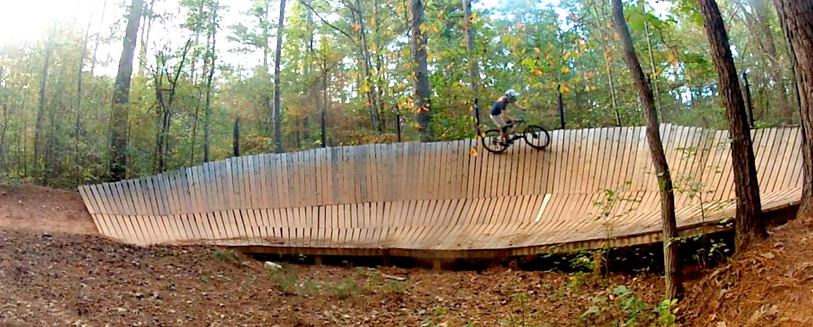 A mountain biker rides along a wooden pump track in a forested area, surrounded by trees with autumn foliage. The track features a curved, ramp-like design, allowing for dynamic movement and skillful riding. Soft dirt and fallen leaves cover the ground below. Blankets Creek mountain bike trail.
