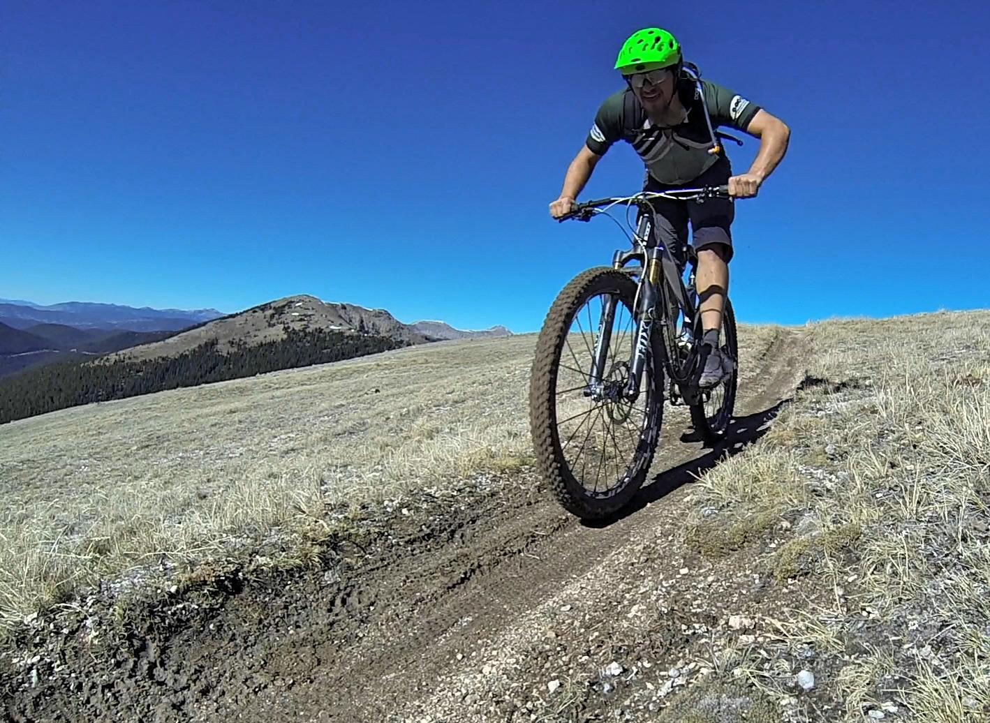 A person riding a mountain bike on a dirt trail in a mountainous landscape, wearing a green helmet and athletic gear, with clear blue skies and distant peaks in the background. Monarch Crest Trail mountain bike trail.