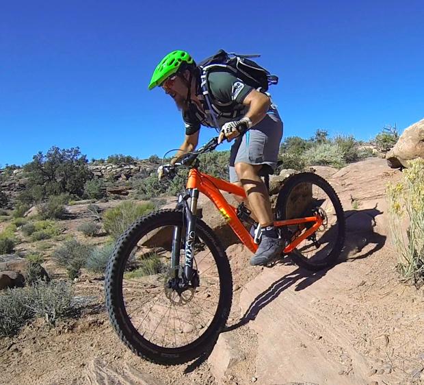 A mountain biker wearing a green helmet and a backpack rides an orange bike along a rocky trail surrounded by shrubs and desert vegetation under a clear blue sky. Arth